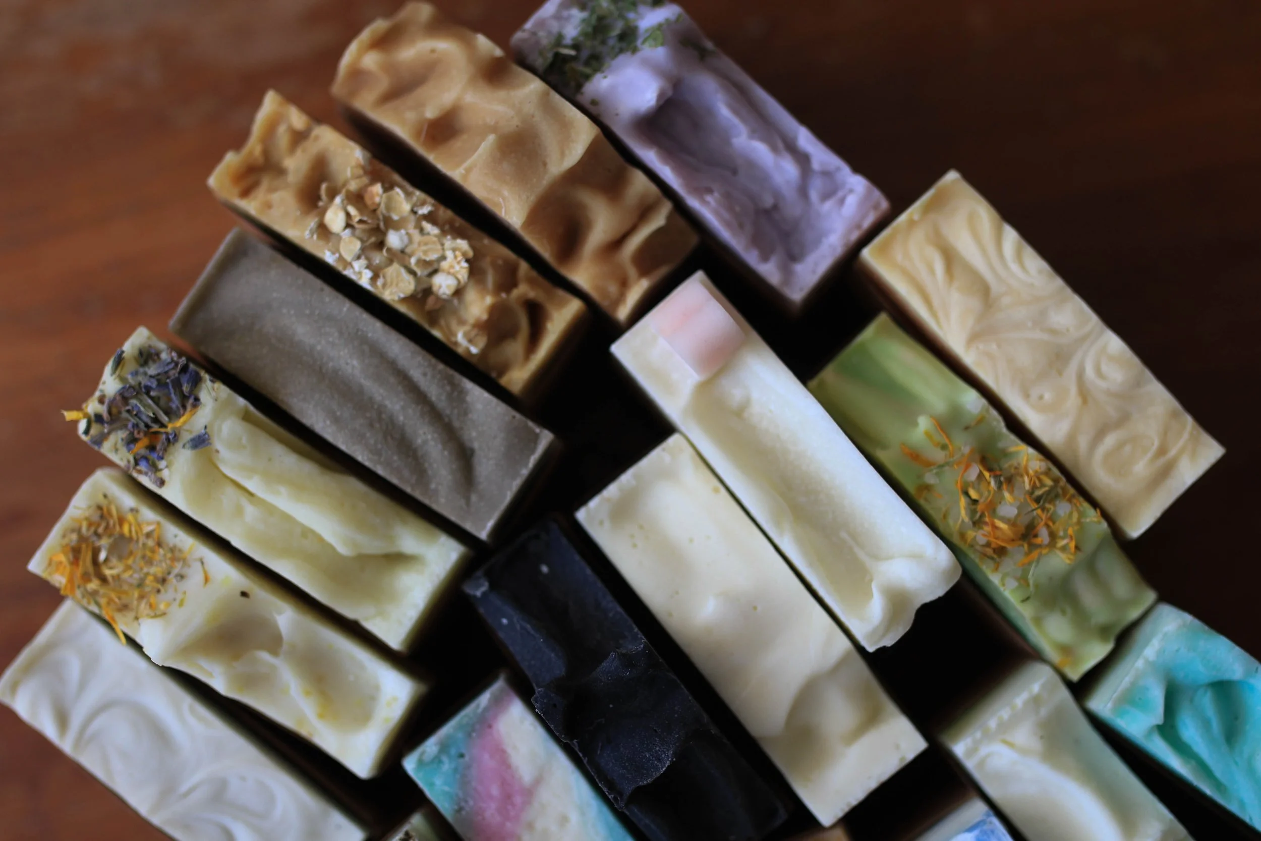 A close-up image of assorted handmade soap bars arranged in a display. The soap bars vary in color, design, and toppings such as dried flowers, herbs, and swirls.