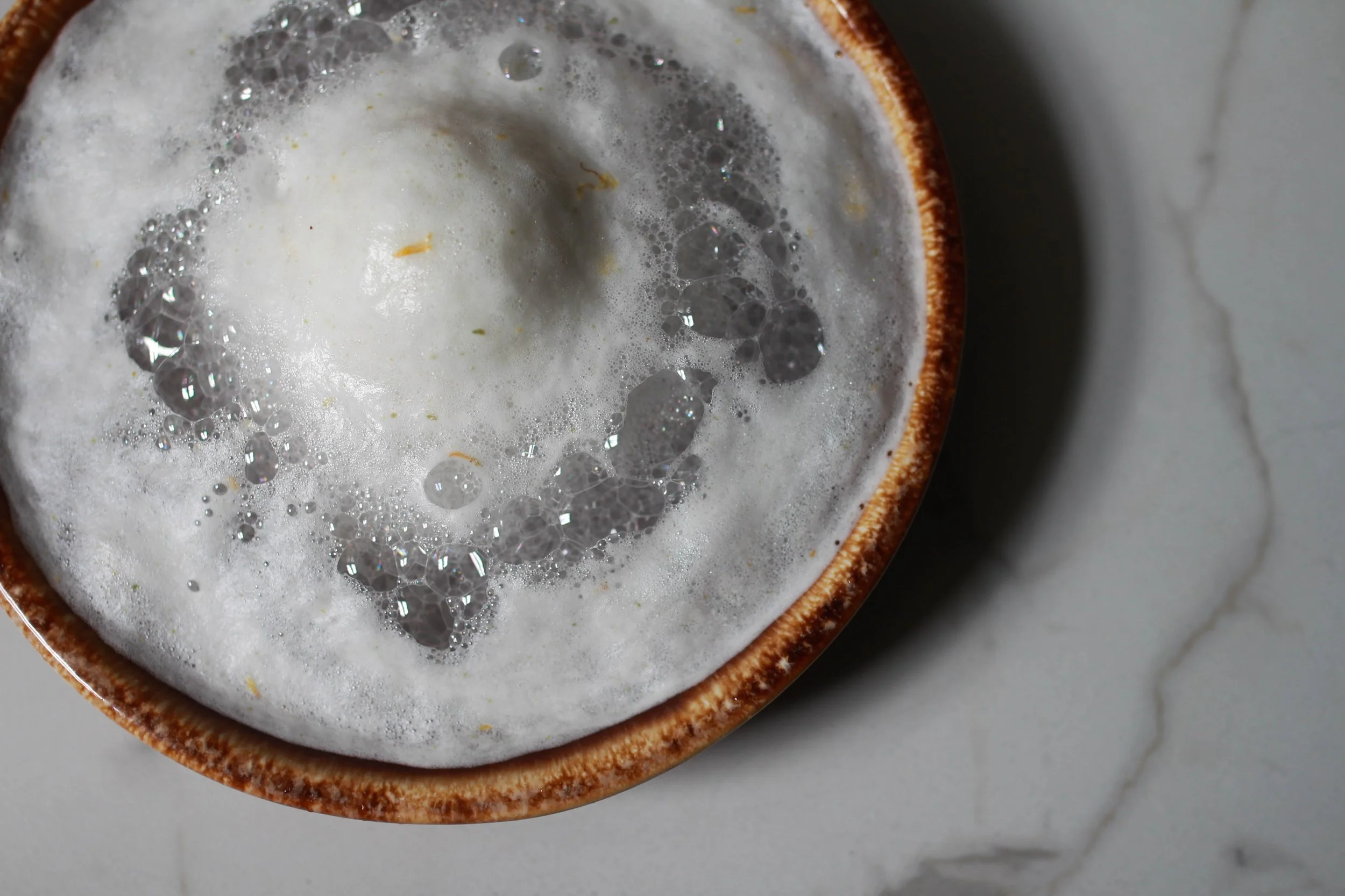 Close-up of a frothy beverage in a brown ceramic cup on a white marble surface.
