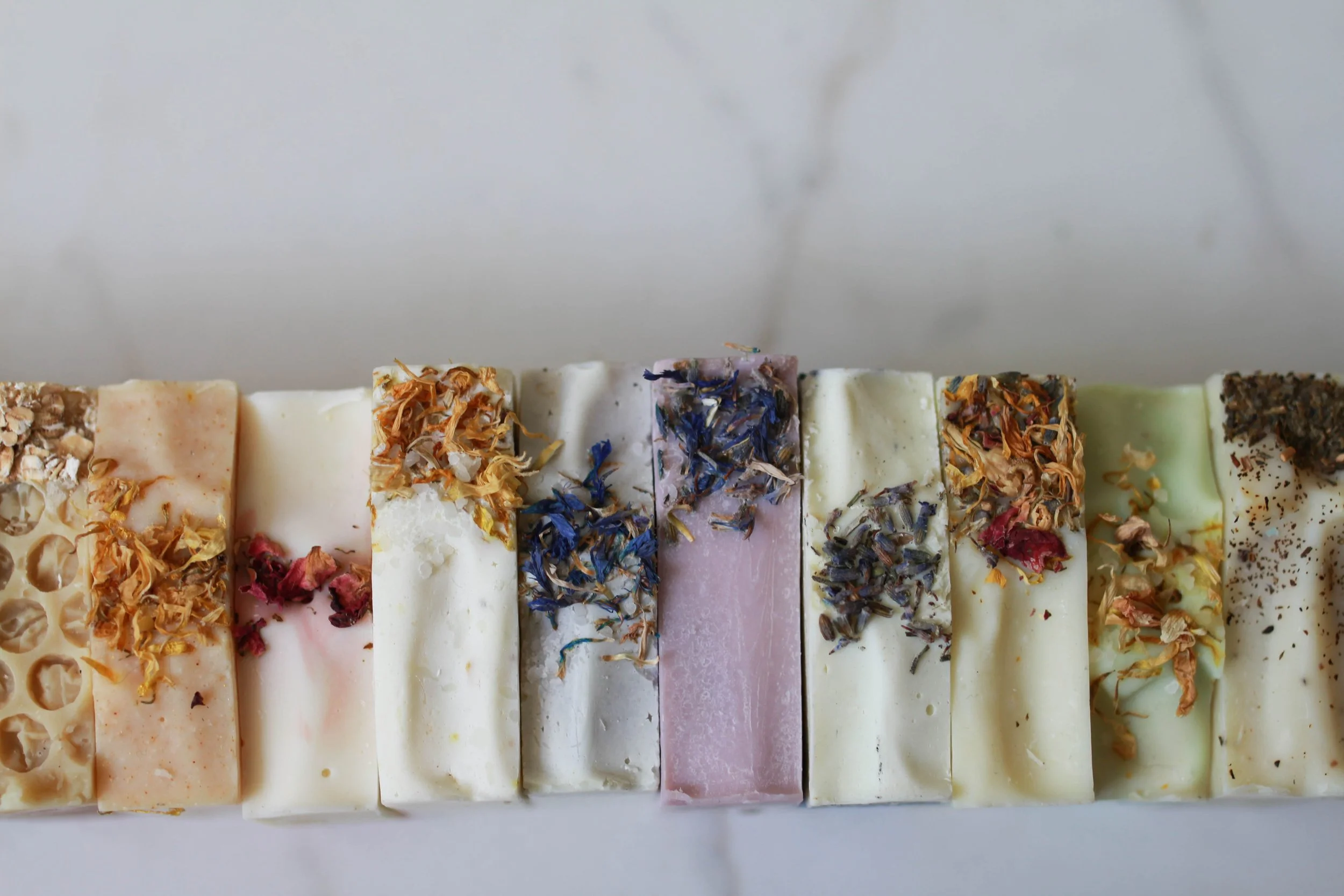 A row of colorful handmade soap bars with various natural ingredients on top, arranged on a white surface.