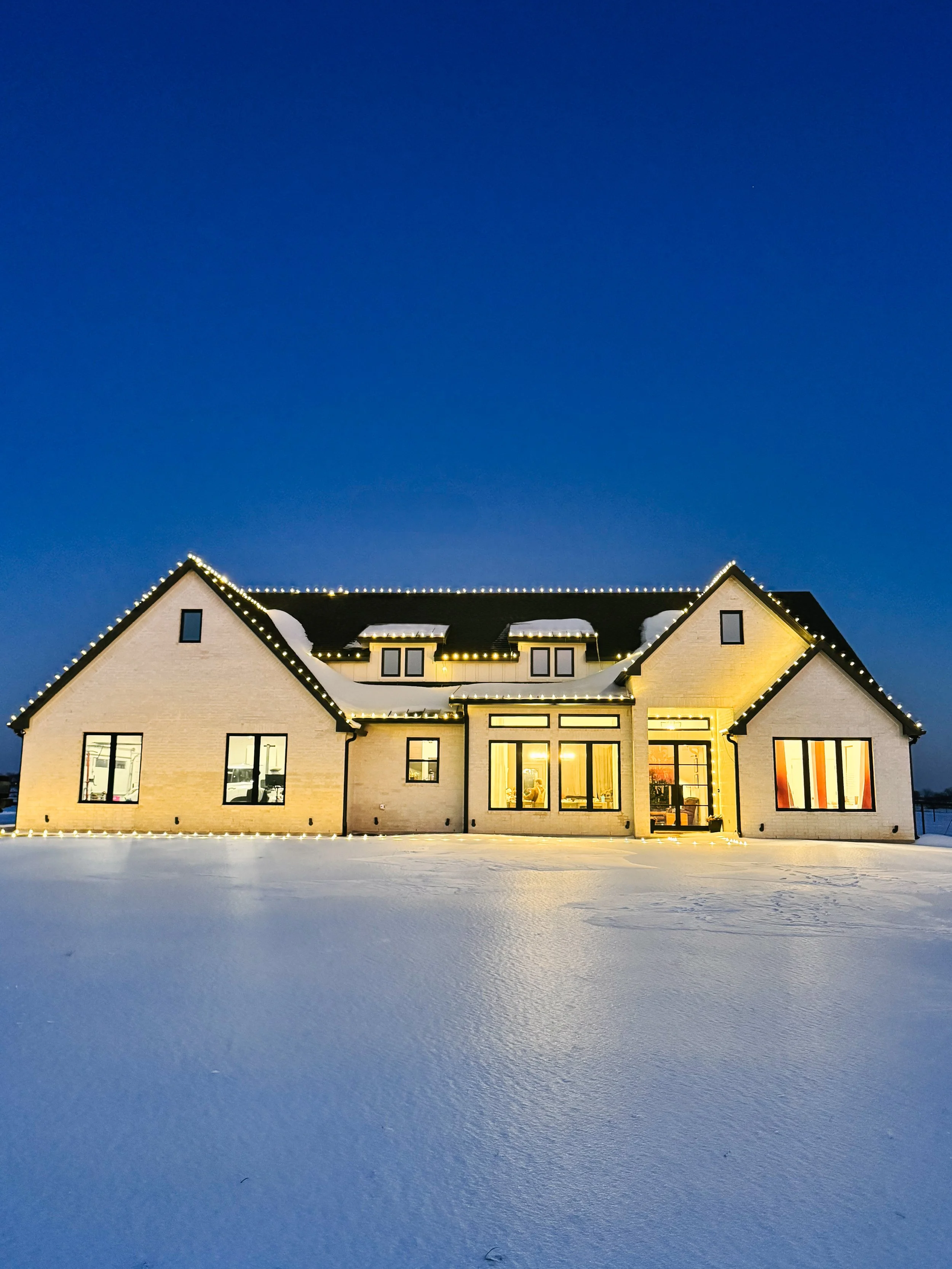 A large house illuminated with Christmas lights at night, surrounded by snow, with a clear dark blue sky overhead.
