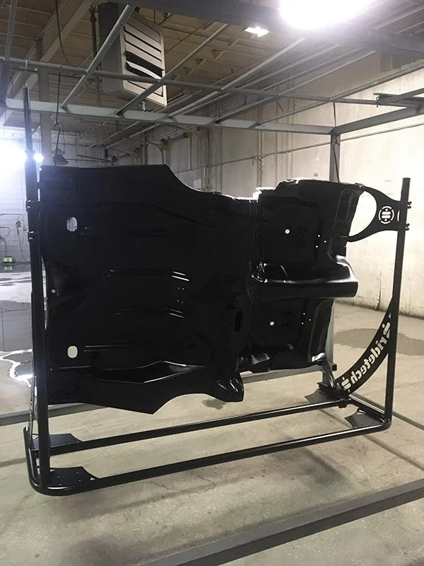 Black powdered coated car chassis or body shell on a metal stand in a warehouse or workshop.