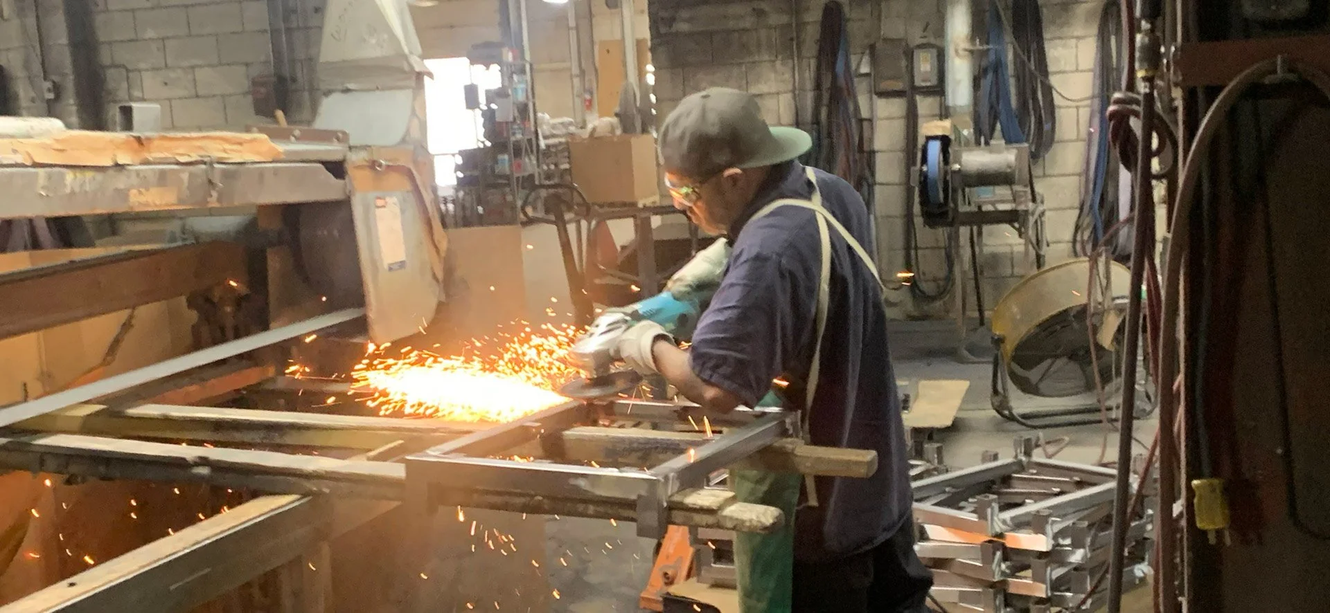 A person working on an industrial metal fabrication project, using a welding machine that produces sparks, in a workshop filled with equipment and supplies.