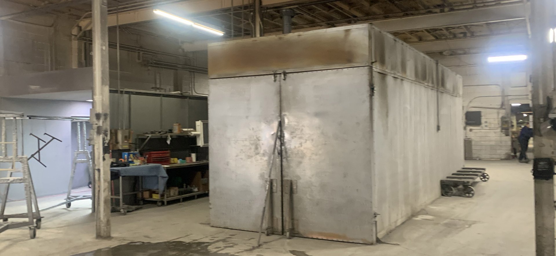 Industrial warehouse with metal storage room, workbenches, and equipment, illuminated by overhead fluorescent lights.