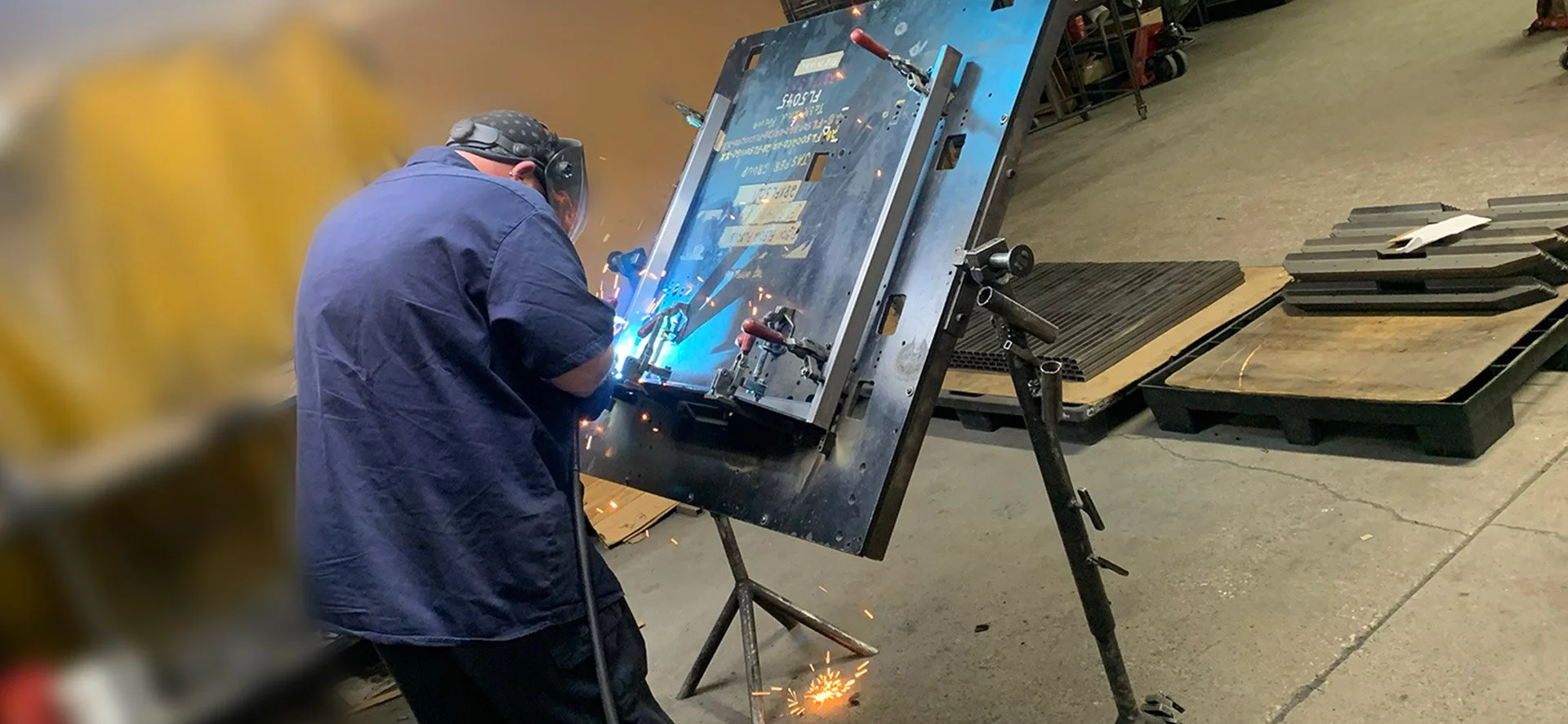 A person welding a metal piece on a large metal structure in a workshop, with sparks flying and protective gear, including a welding helmet.