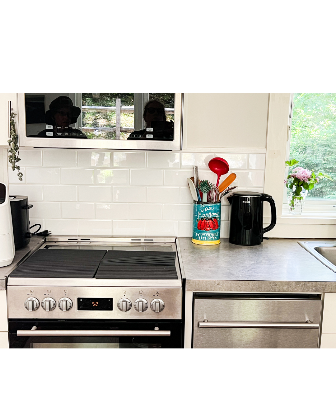 Modern kitchen countertop with electric stove, black electric kettle, hand-made ceramic utensil holder with red spoon, orange spatula, white spatula, and green cleaning brush, white tiled backsplash, window with pink flowers outside.