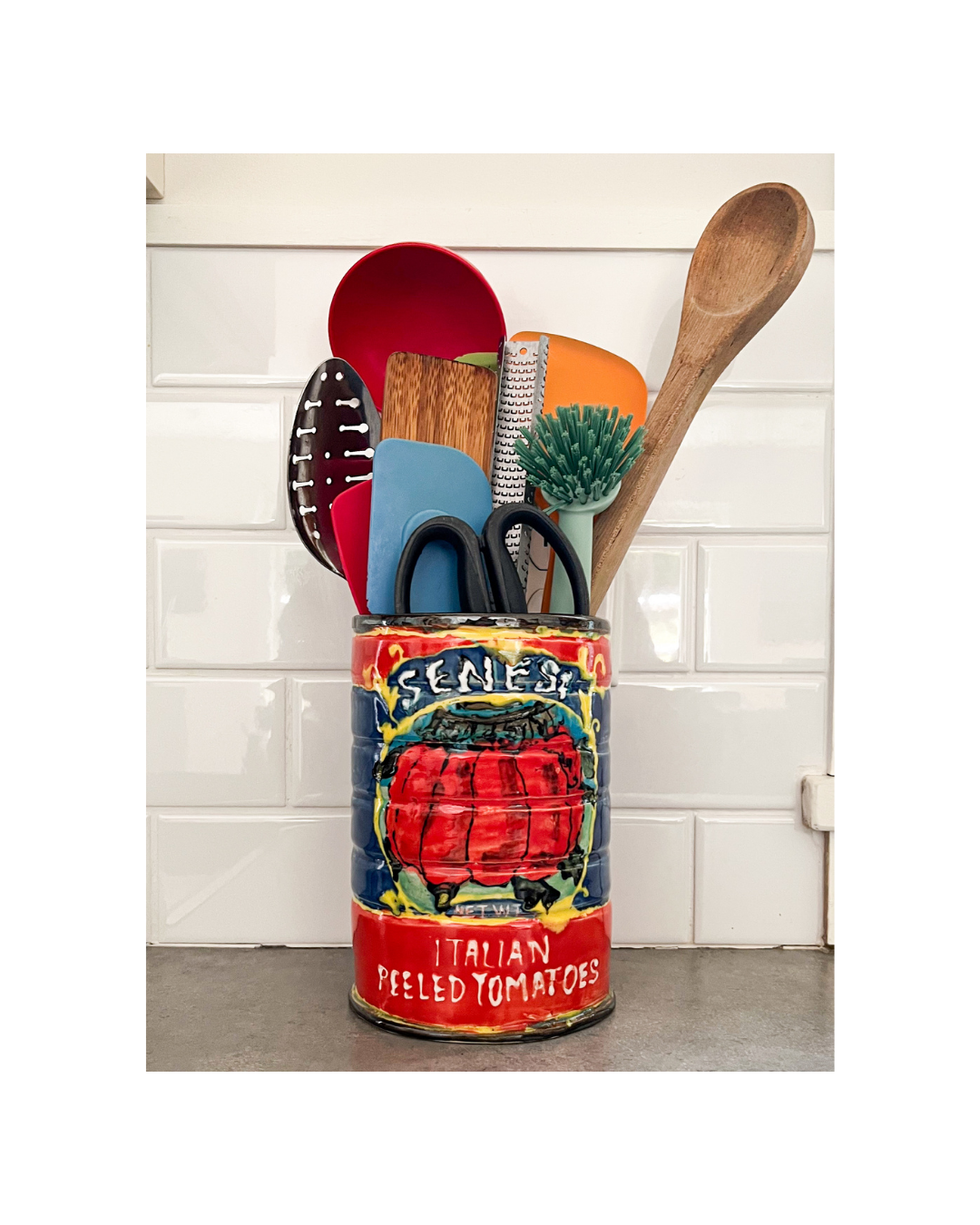 Collection of kitchen utensils in a colorful painted ceramic can on a kitchen countertop, including wooden spoons, spatulas, a vegetable brush, and scissors, against a white tiled backsplash.