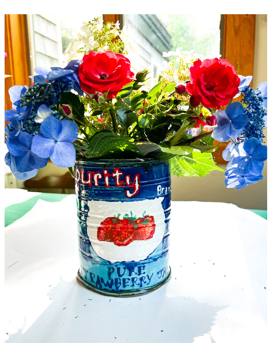 A colorful bouquet of flowers including red roses, blue hydrangeas, and white filler flowers in a hand painted ceramic can with strawberry and raspberry illustrations, placed on a white surface near a window.