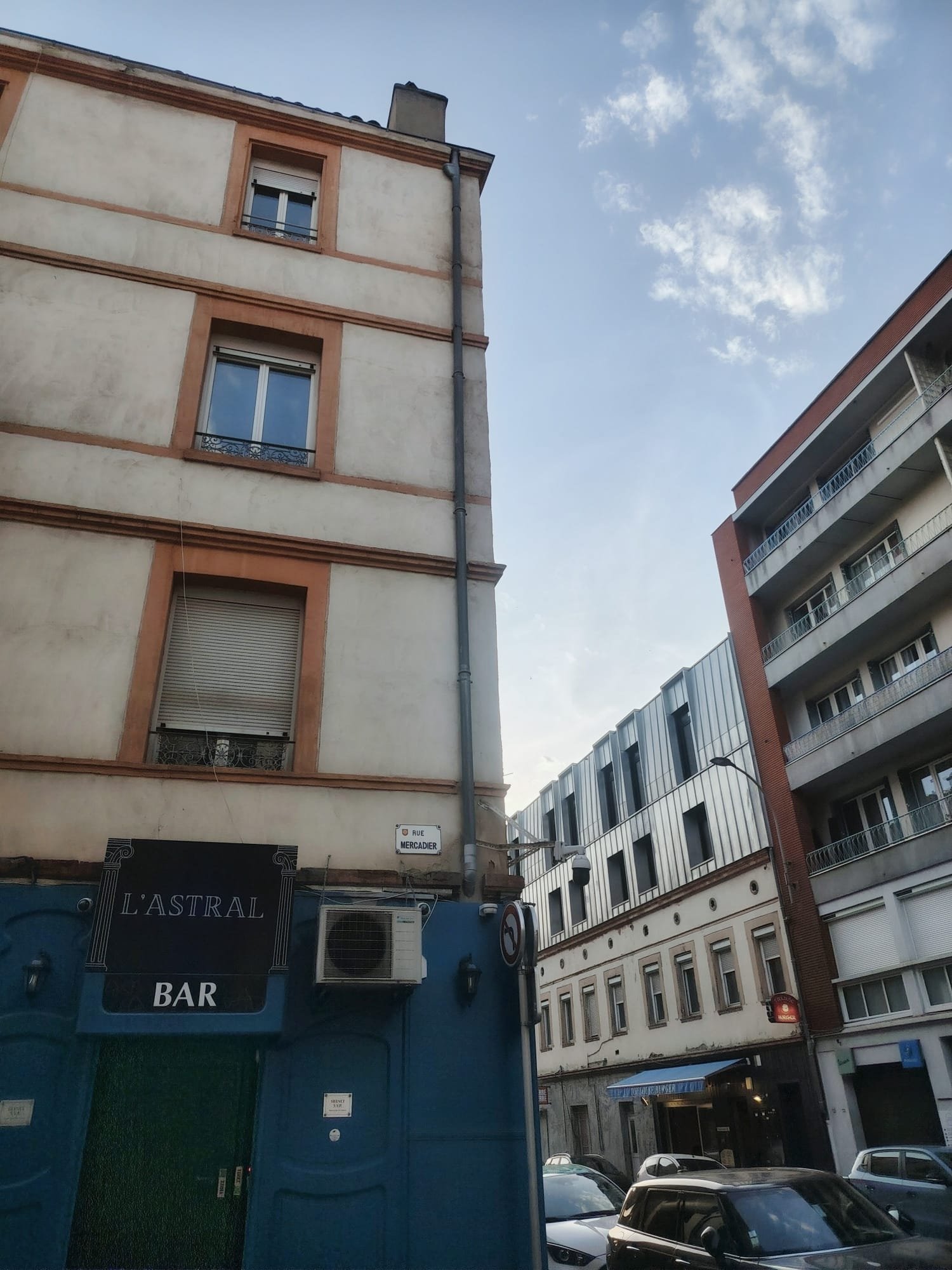 A city street scene with a blue bar called L'Astral, multi-story residential buildings, cars parked along the street, and a street sign that reads Rue Mercadier. The sky is partly cloudy.