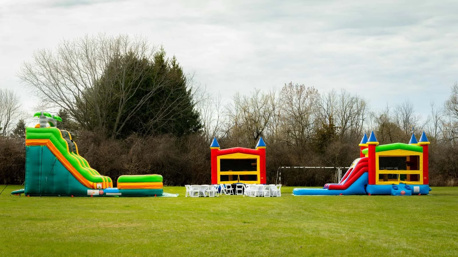 Two colorful inflatable bounce houses shaped like castles with slides are set up on a grassy field, with a table and chairs in the middle and a backdrop of trees under a cloudy sky.