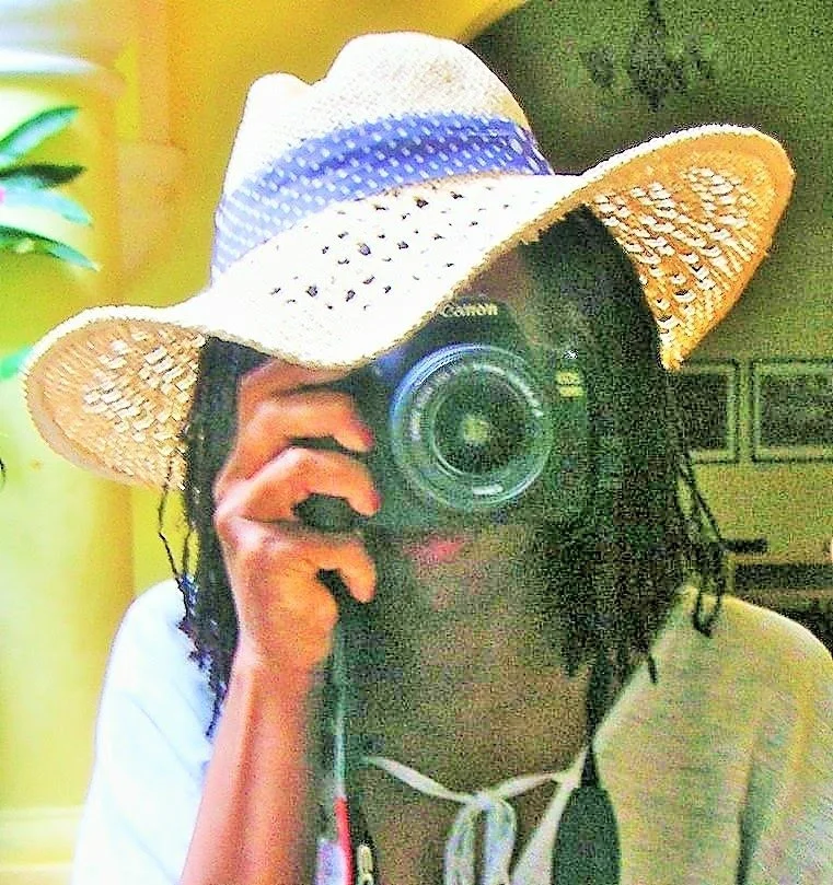 Person taking a photo with a camera, wearing a large straw hat and a white hoodie, indoors with green and yellow walls.