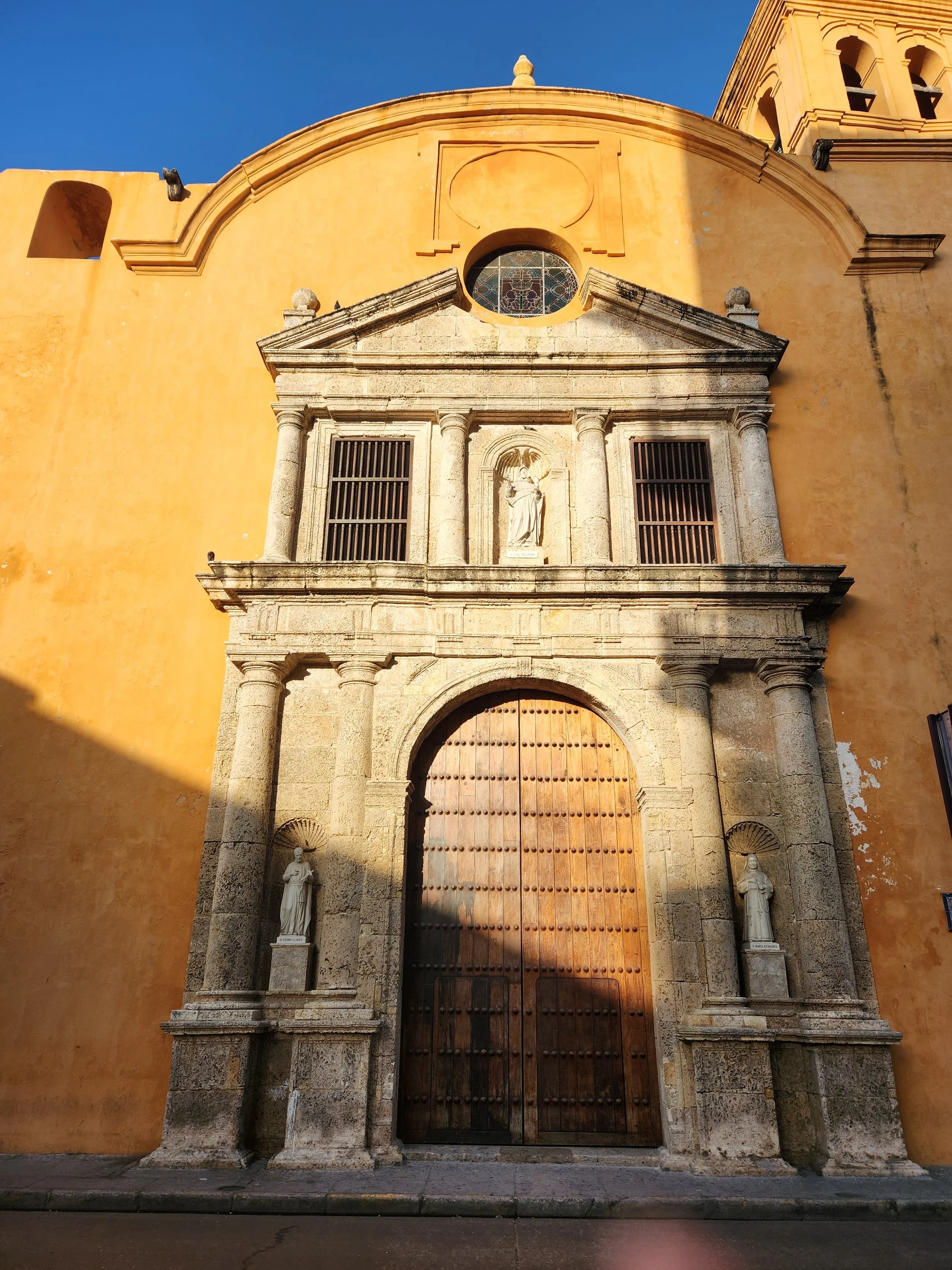 Facade of a historic church with a large wooden door, stone columns, statues on pedestals, and a small round stained glass window at the top, painted in warm yellow tones under a clear blue sky.