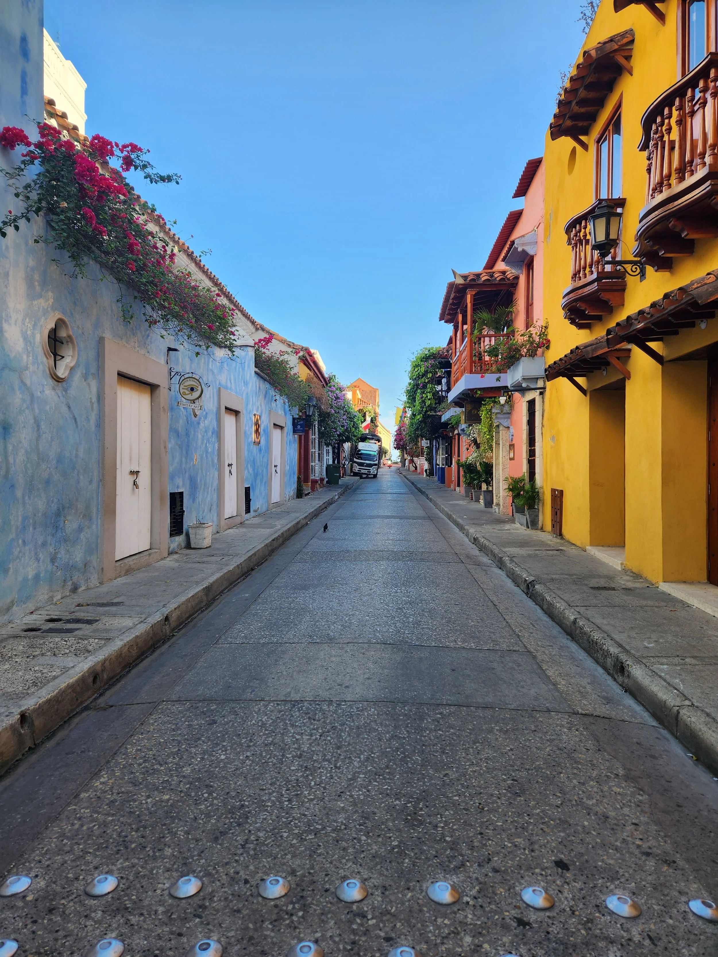 A colorful narrow street with pastel yellow and blue buildings, flowered balconies, and a clear blue sky.