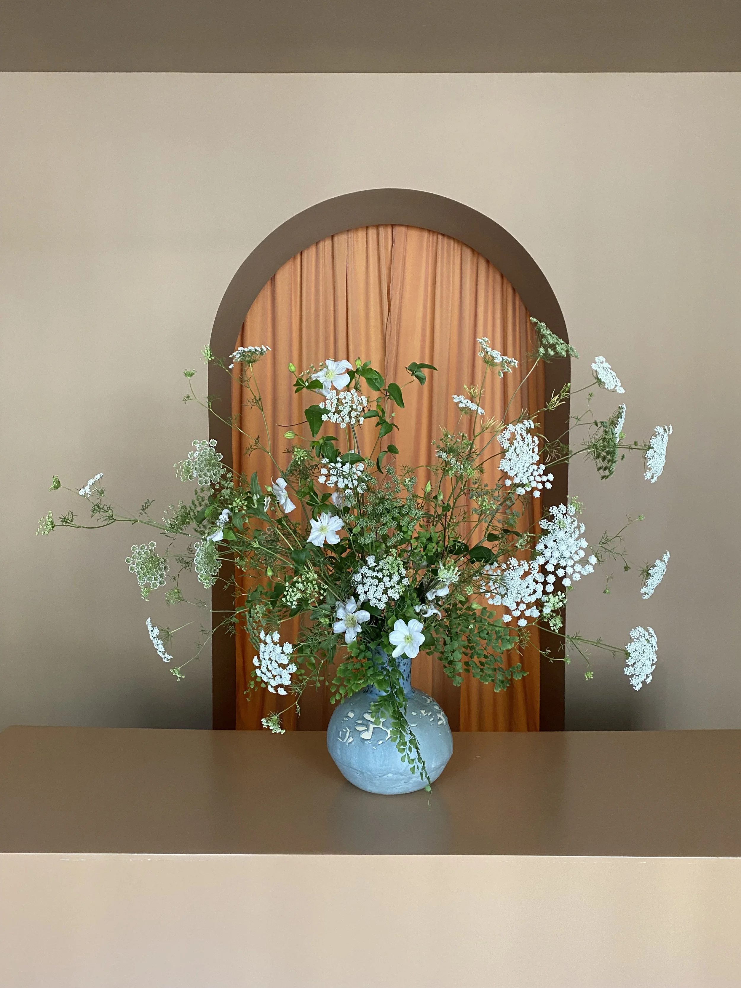 A ceramic vase filled with white flowers and green foliage sits on a beige surface, in front of a wooden door with an arched top, in an indoor setting.