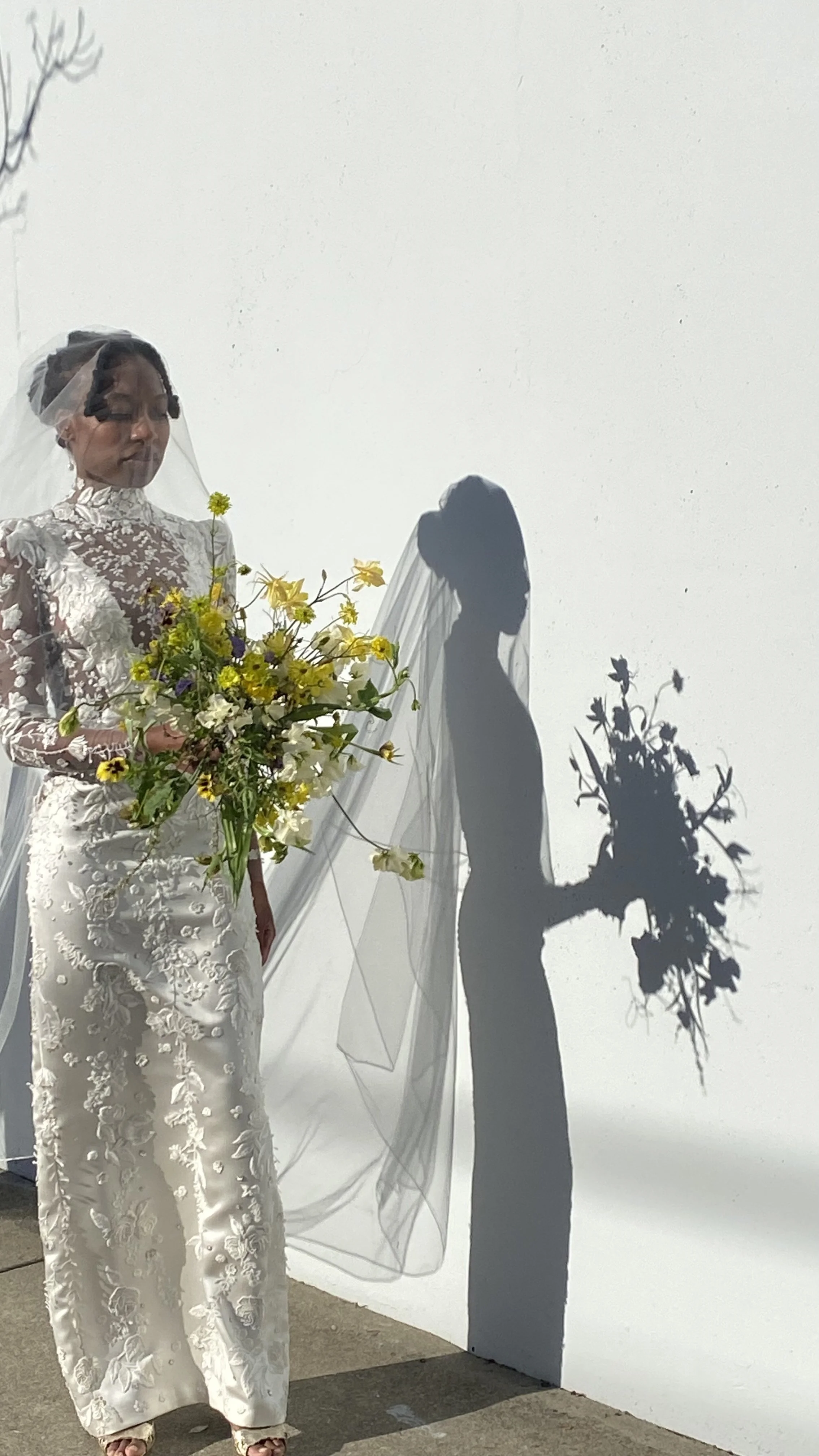 A bride in a wedding dress holding a bouquet of flowers, casting a shadow on a plain white wall.