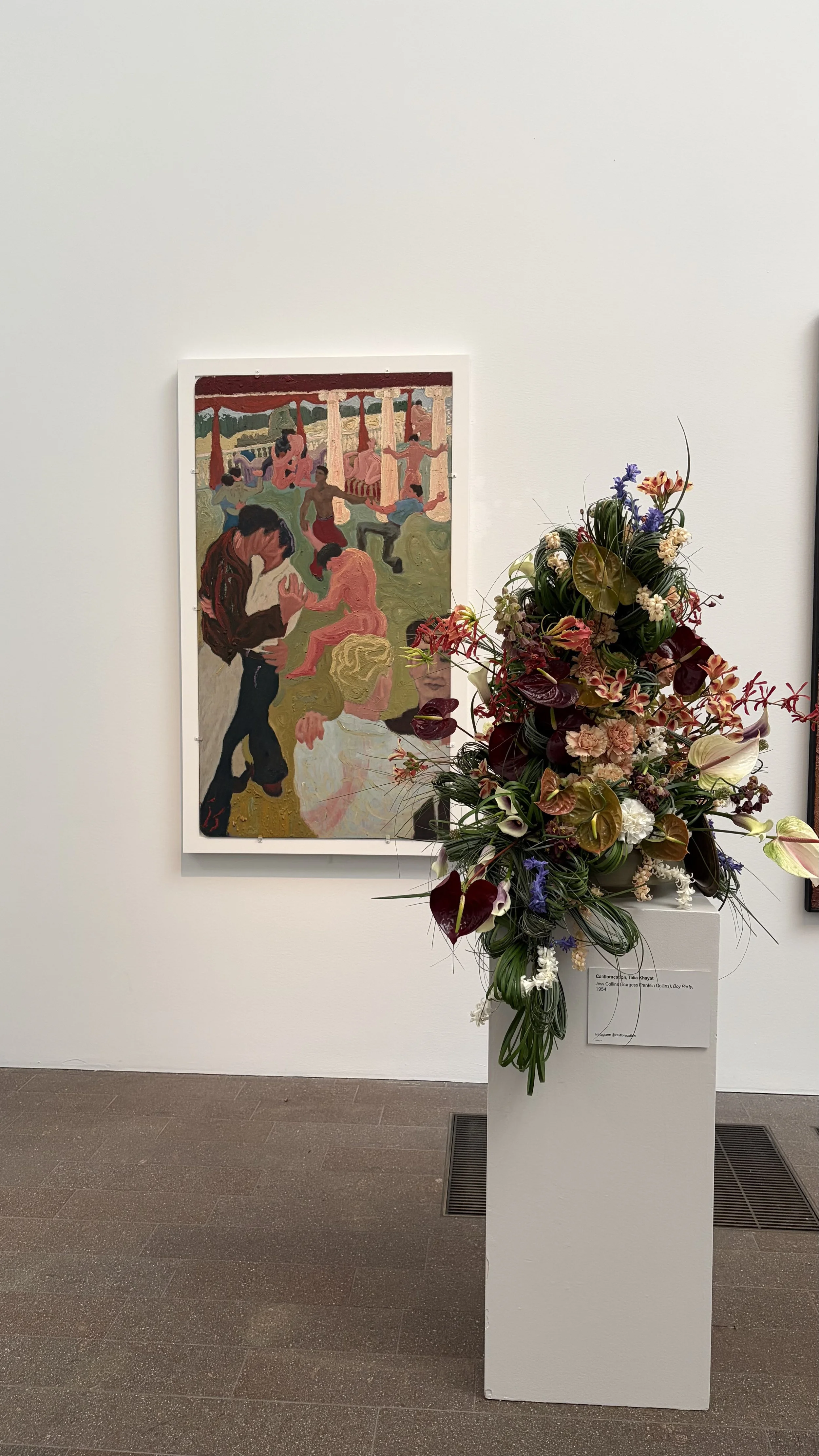 A colorful floral arrangement on a white pedestal in front of a painting of dancers, in the deYoung Museum in San Francisco
