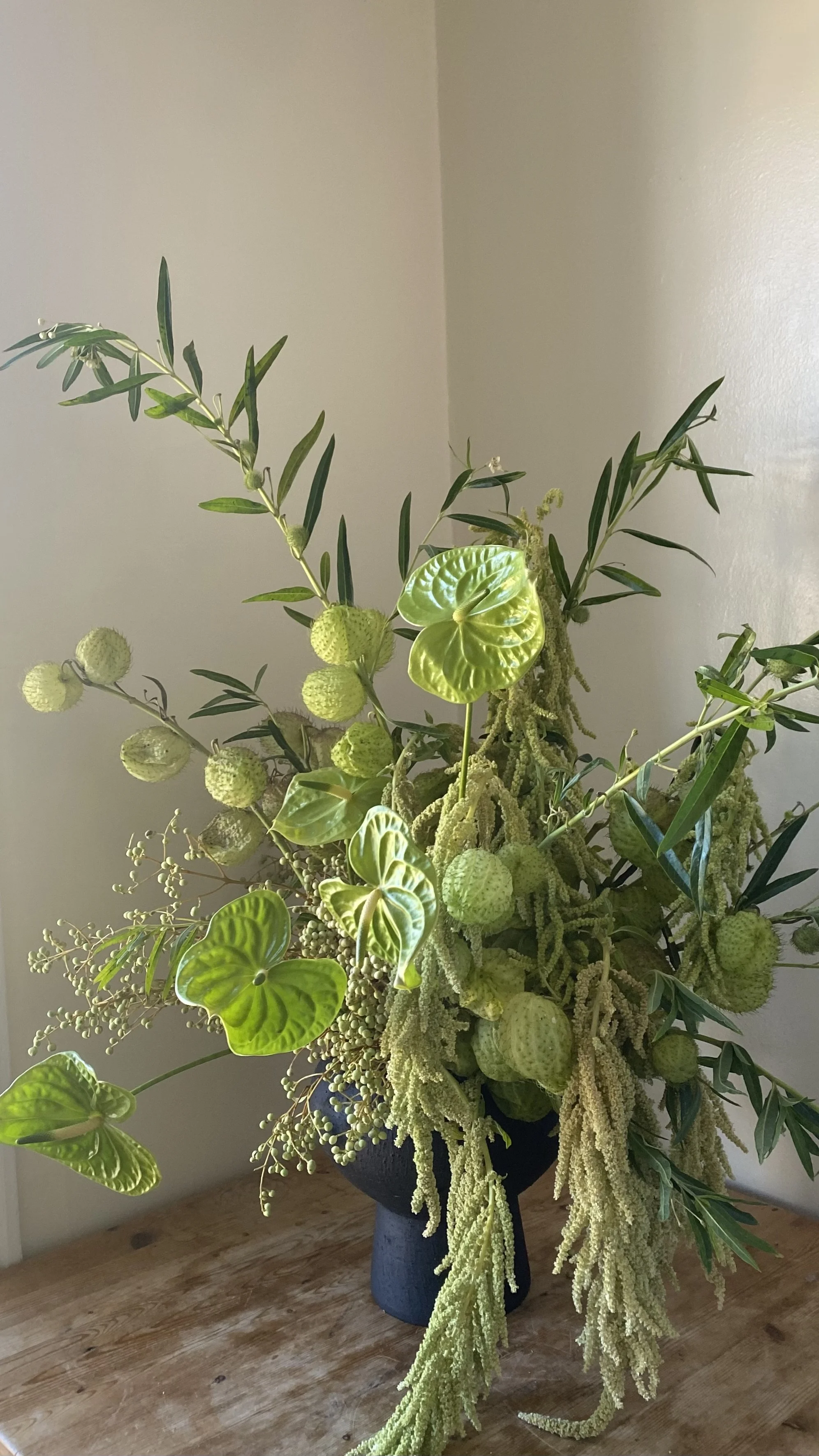 A floral arrangement in a black vase with various greenery and plant varieties, including large green leaves, hanging yellowish-green flowers, and long trailing stems, placed on a wooden surface against a plain beige wall.