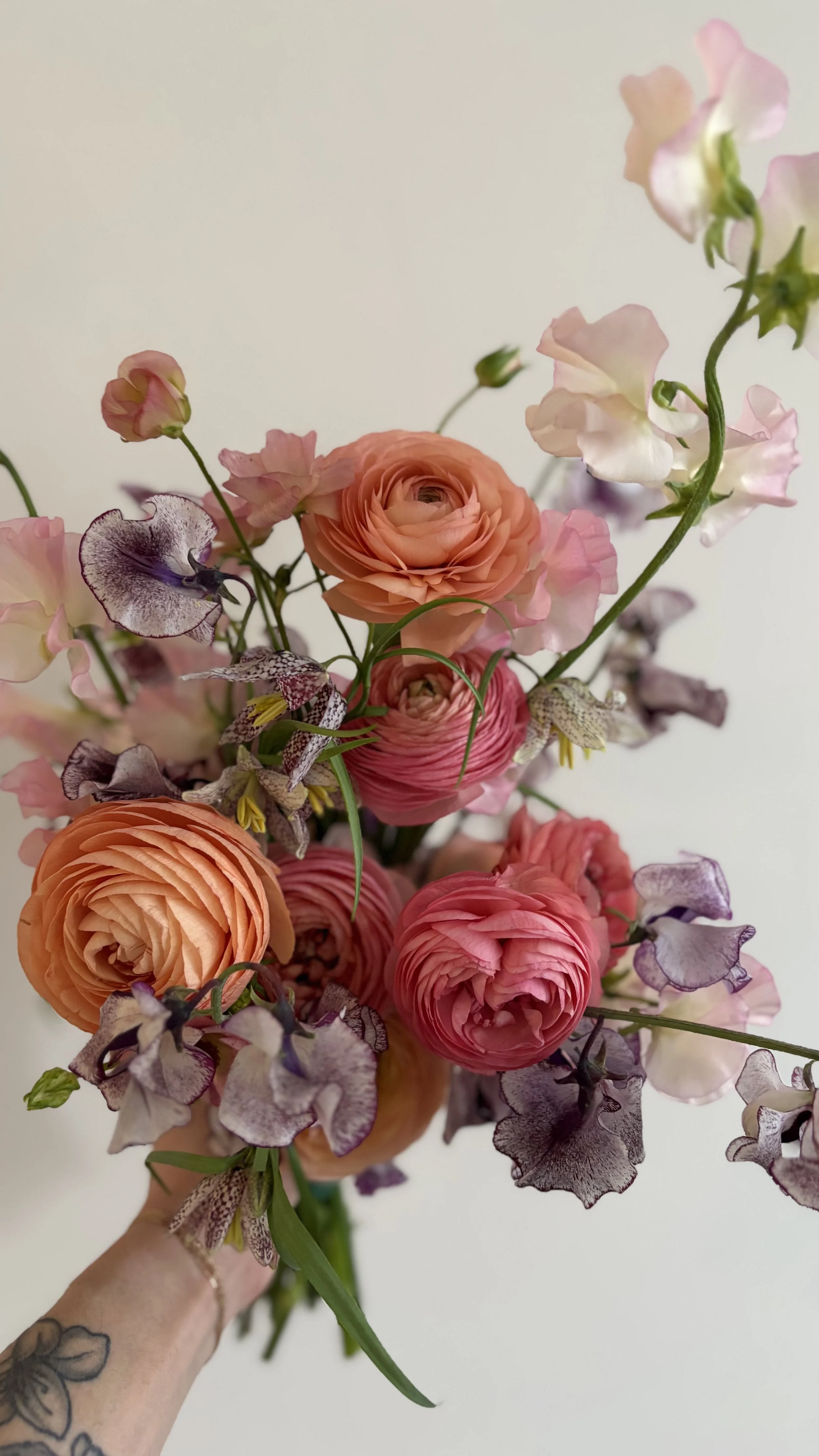 A person holding a bouquet of pink, peach, and purple flowers against a plain light background.