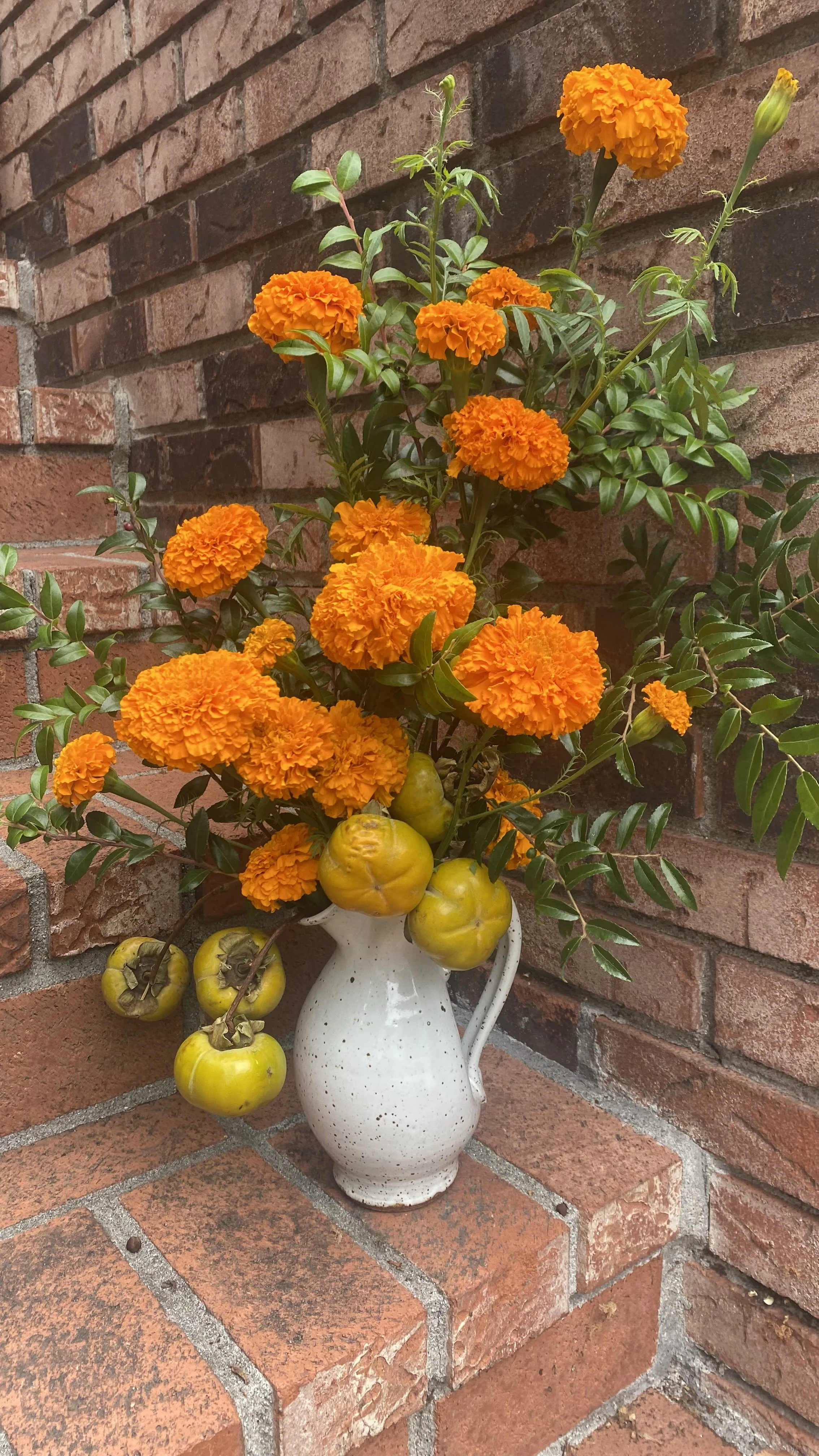 A white speckled ceramic pitcher holding a bouquet of orange marigold flowers and yellow tomatoes, placed on a brick ledge against a brick wall.