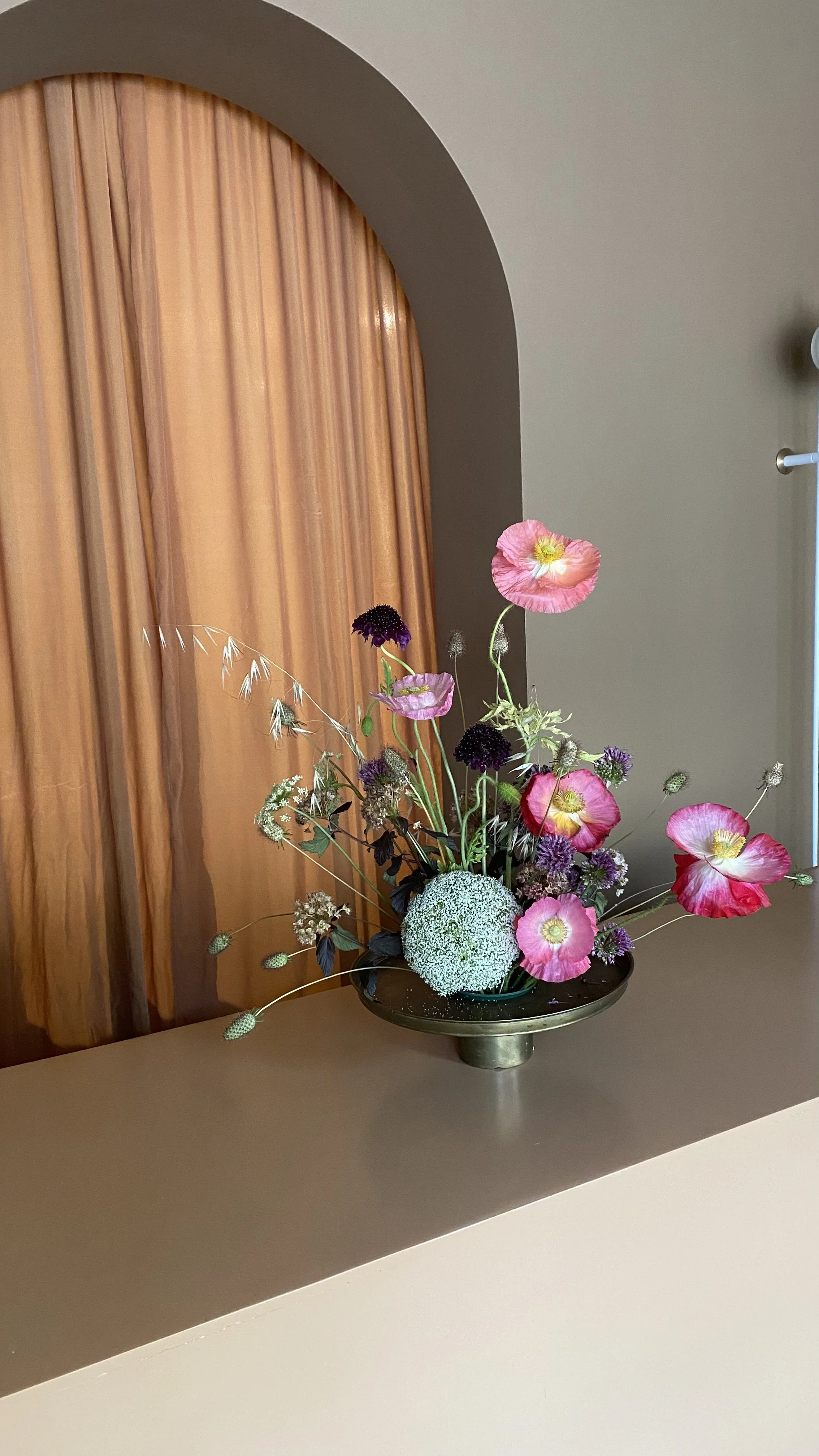 A floral arrangement with pink poppies, purple alliums, and white caps in a black and gold vase on a beige countertop.