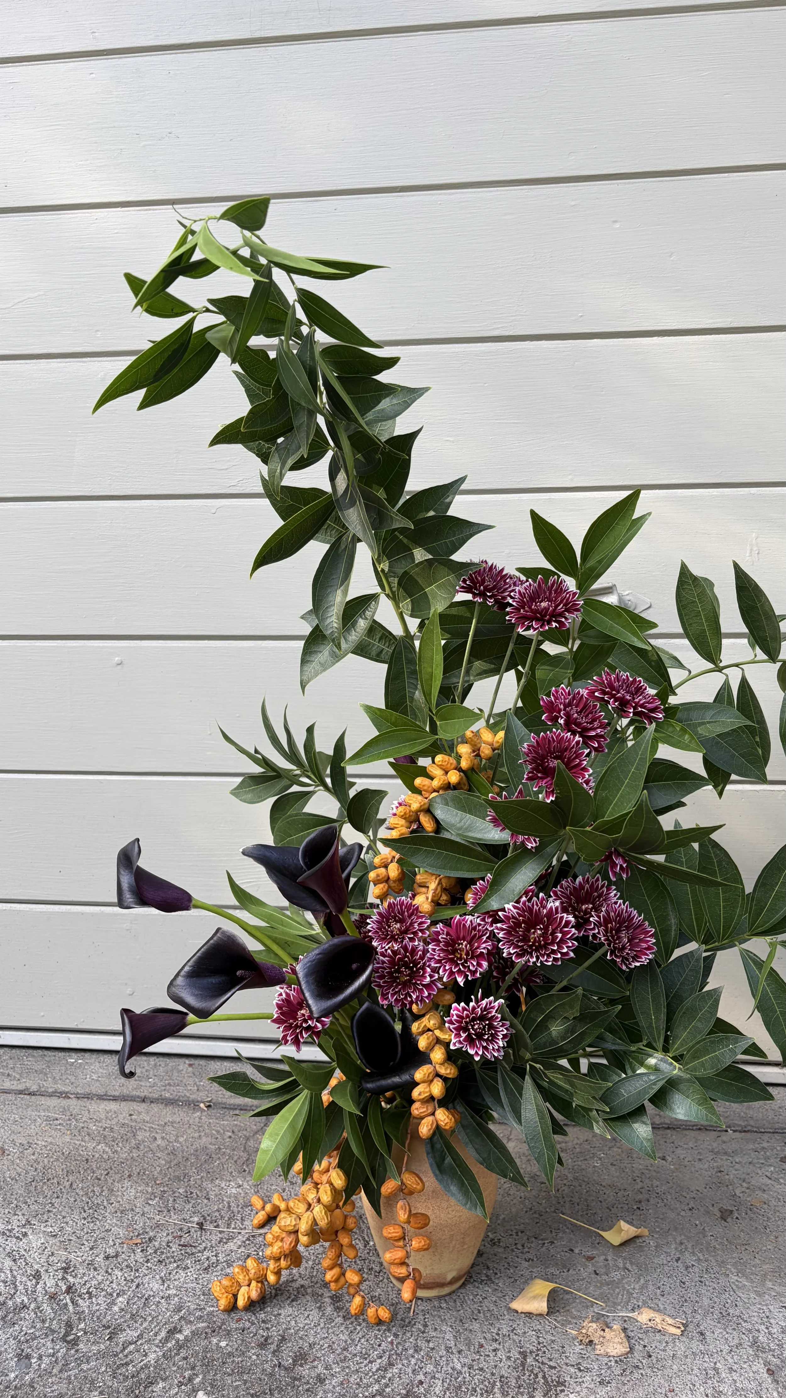 Flower arrangement with purple calla lilies, dark purple flowers, and pink and white chrysanthemums in a beige pot, with green foliage, against a white wooden wall background.