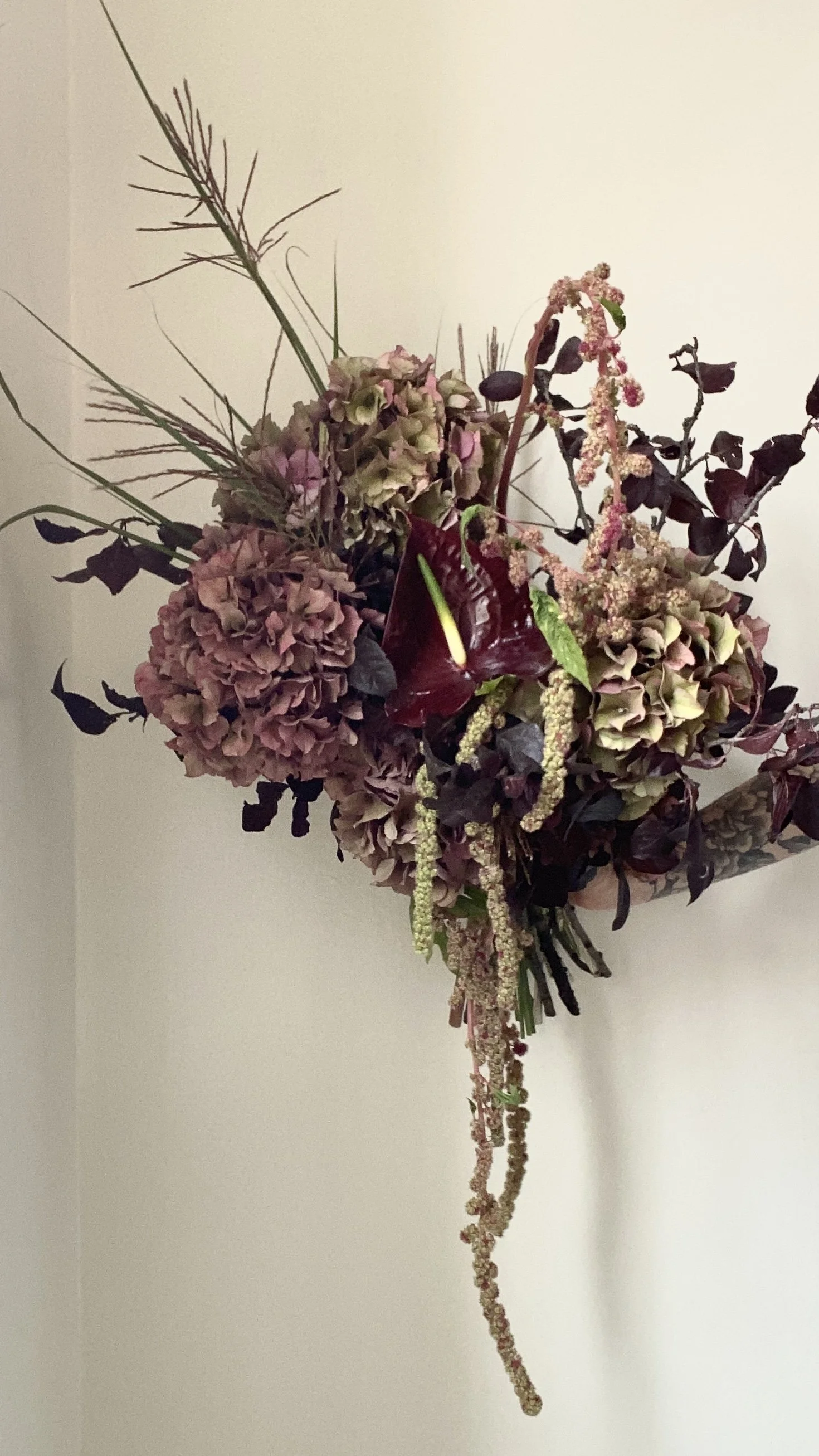 A floral wall arrangement with pink and purple hydrangeas, dark purple leaves, greenery, and hanging amaranthus, mounted on a corner wall.