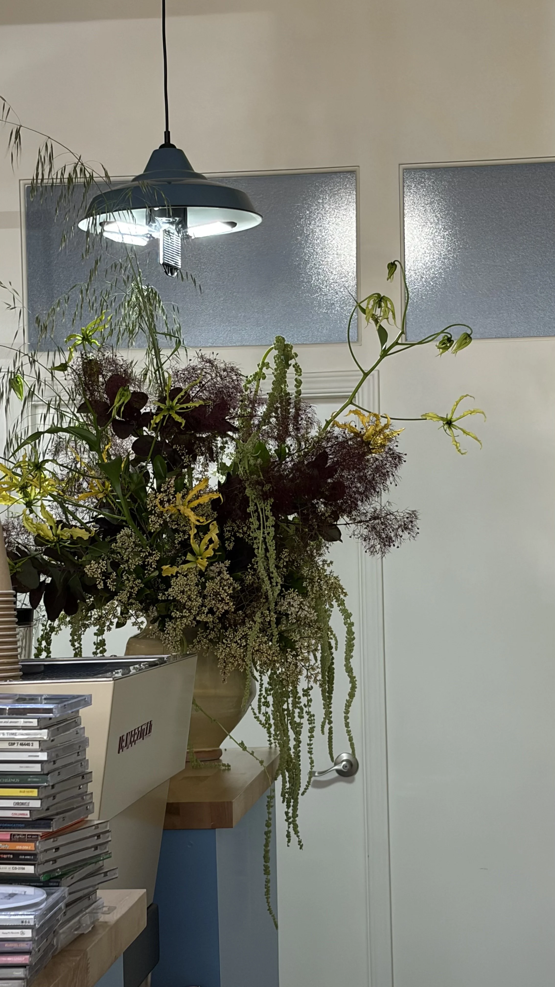 A large flower arrangement with various green and dark  flowers is on a wooden table at Third Wheel Coffee San Francisco. A modern black ceiling light hangs above, and there are two textured glass windows on a white wall in the background.