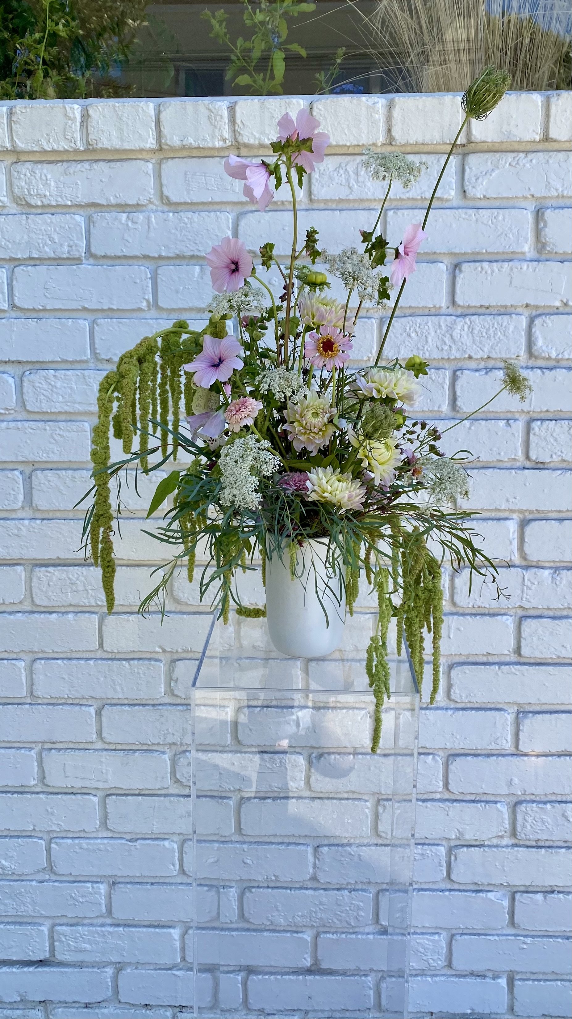 A colorful flower arrangement with various pink and white flowers and green foliage in a white vase, displayed on a clear stand against a white brick wall.