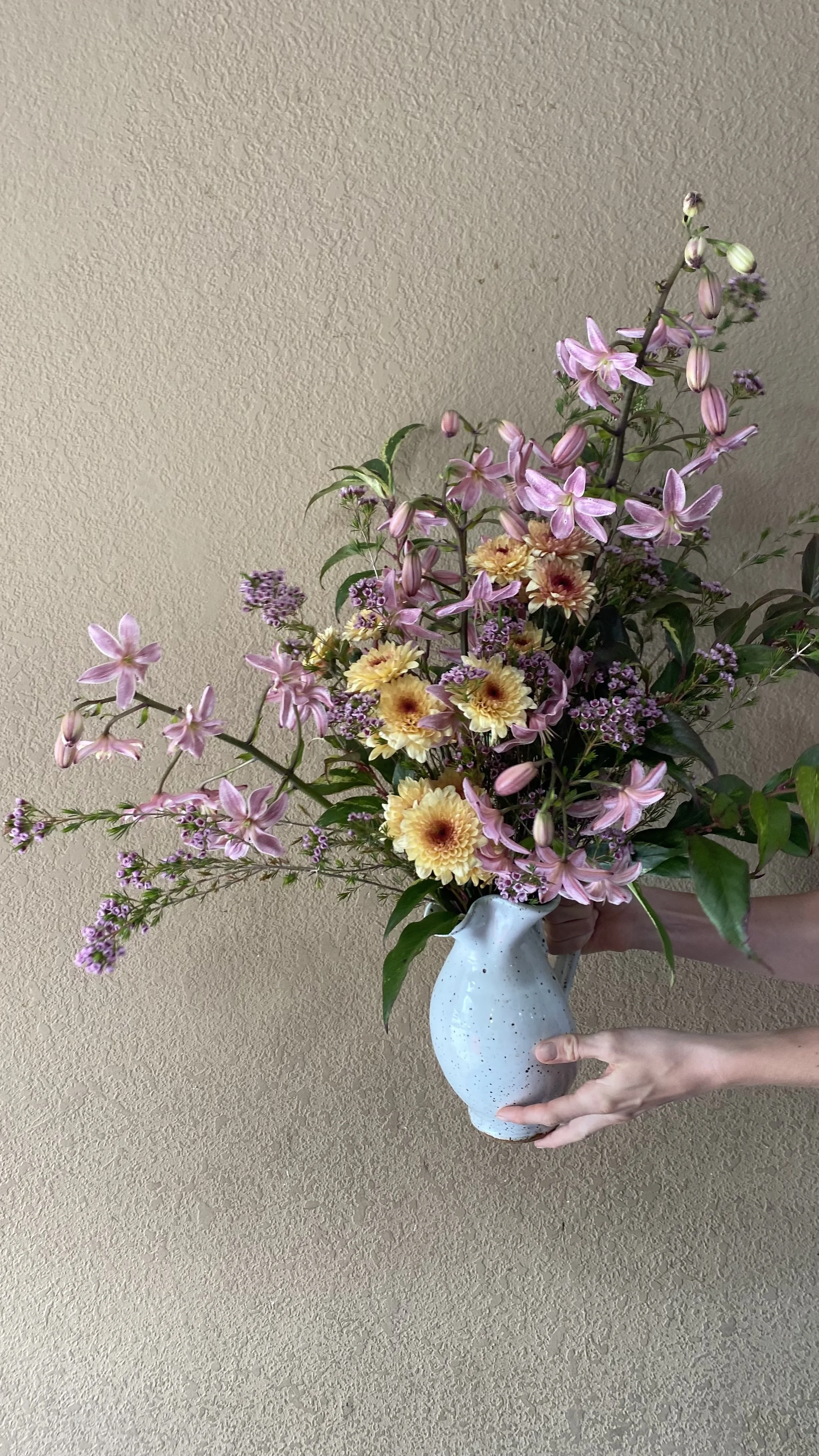 A person holding a tall white ceramic vase filled with pink, purple, and yellow flowers against a beige wall.