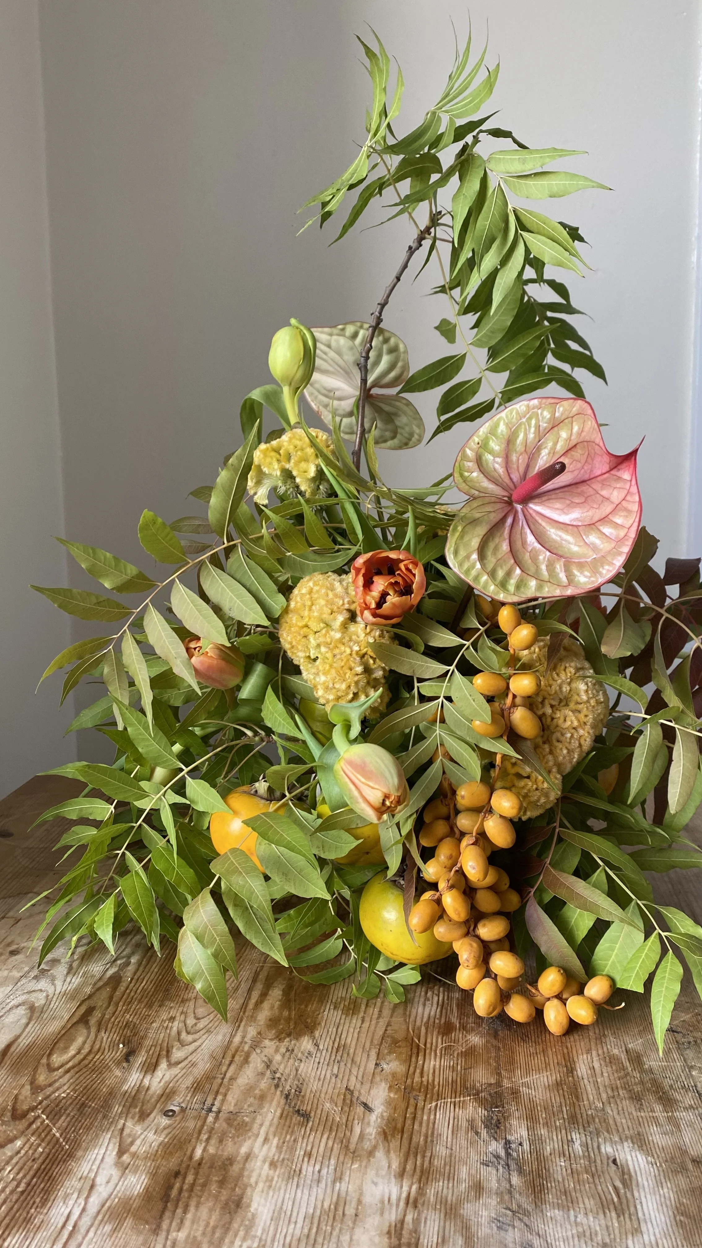 A floral arrangement with various green leaves, pink and yellow flowers, and clusters of yellow and orange berries on a wooden surface.