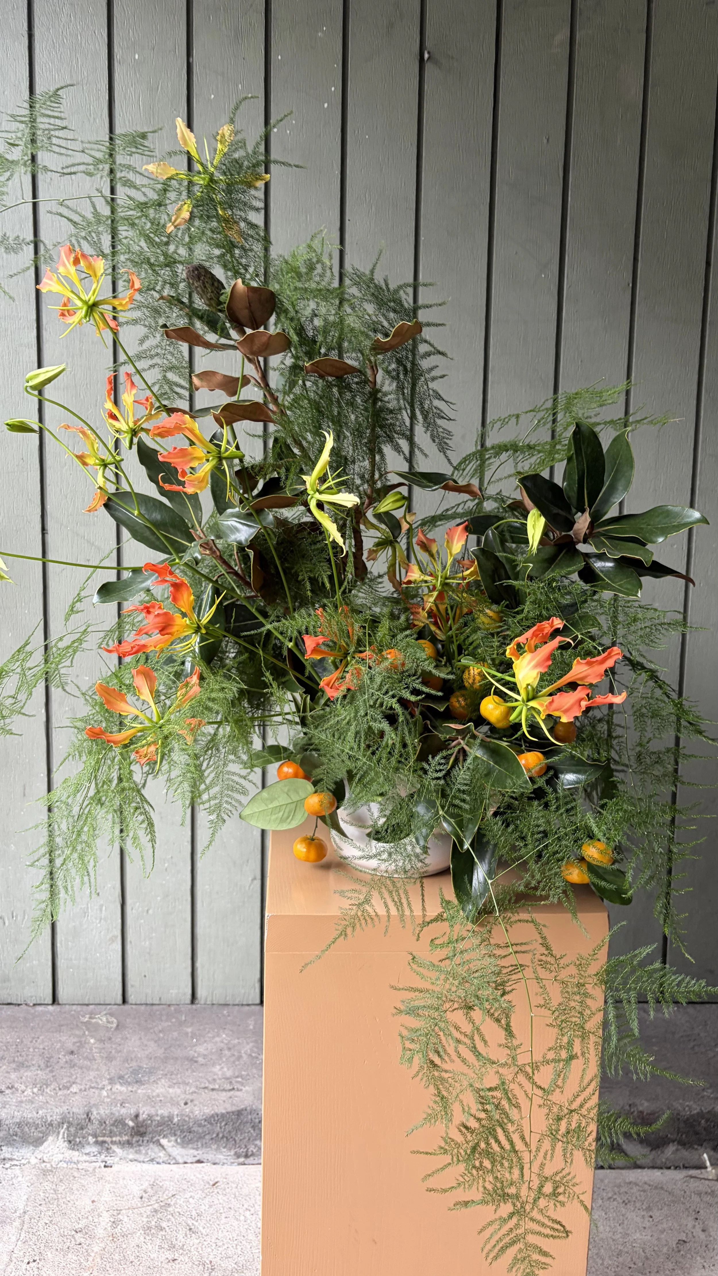 A large flower arrangement with orange and yellow gloriosa lilies, green foliage, and small orange fruits in a white pot on a tan pedestal against a wooden gray wall.