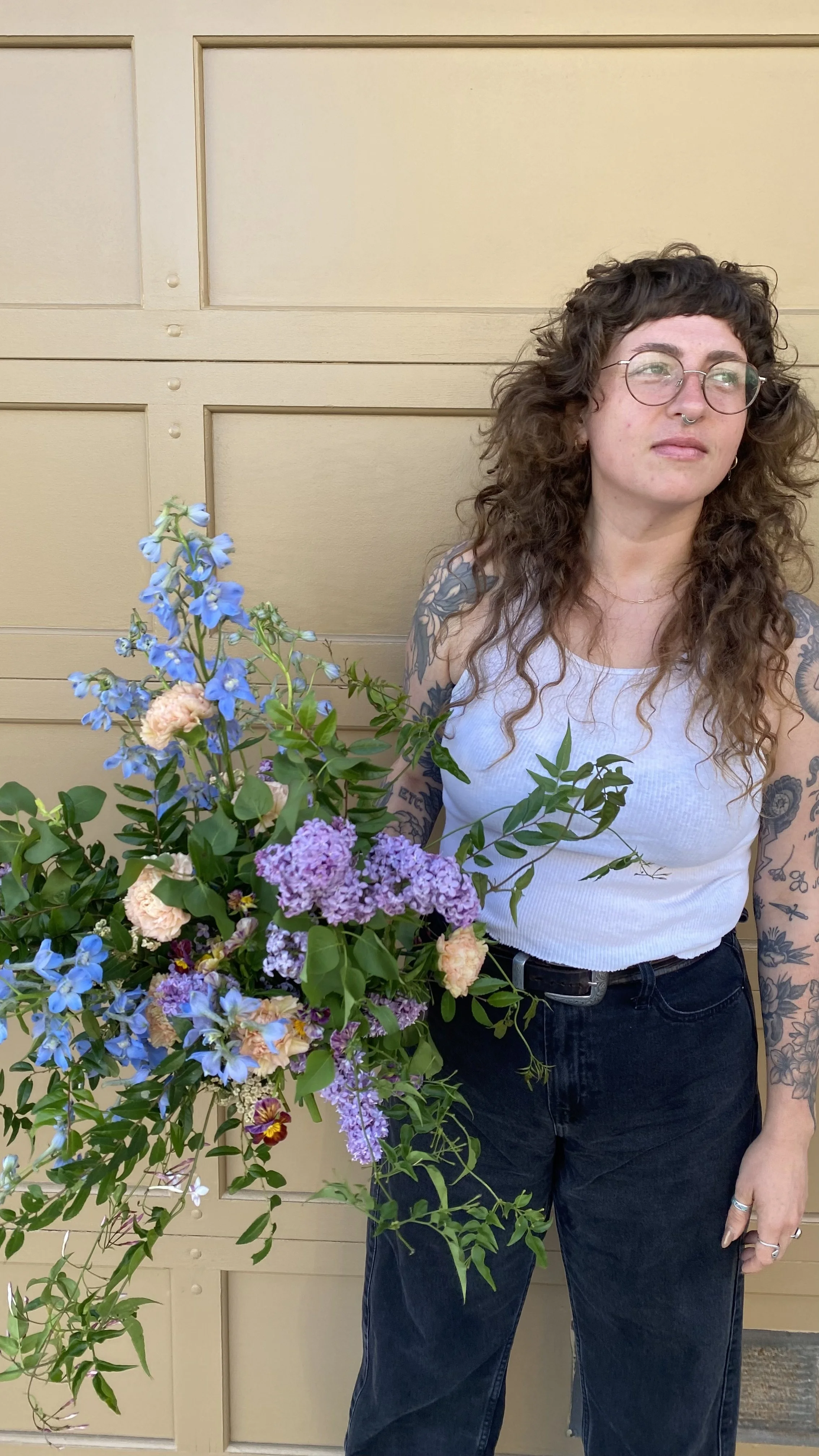A woman with curly brown hair, glasses, a septum piercing, and tattoos on her arms, wearing a white tank top and black jeans, holding a large bouquet of colorful flowers, standing in front of a beige garage door.