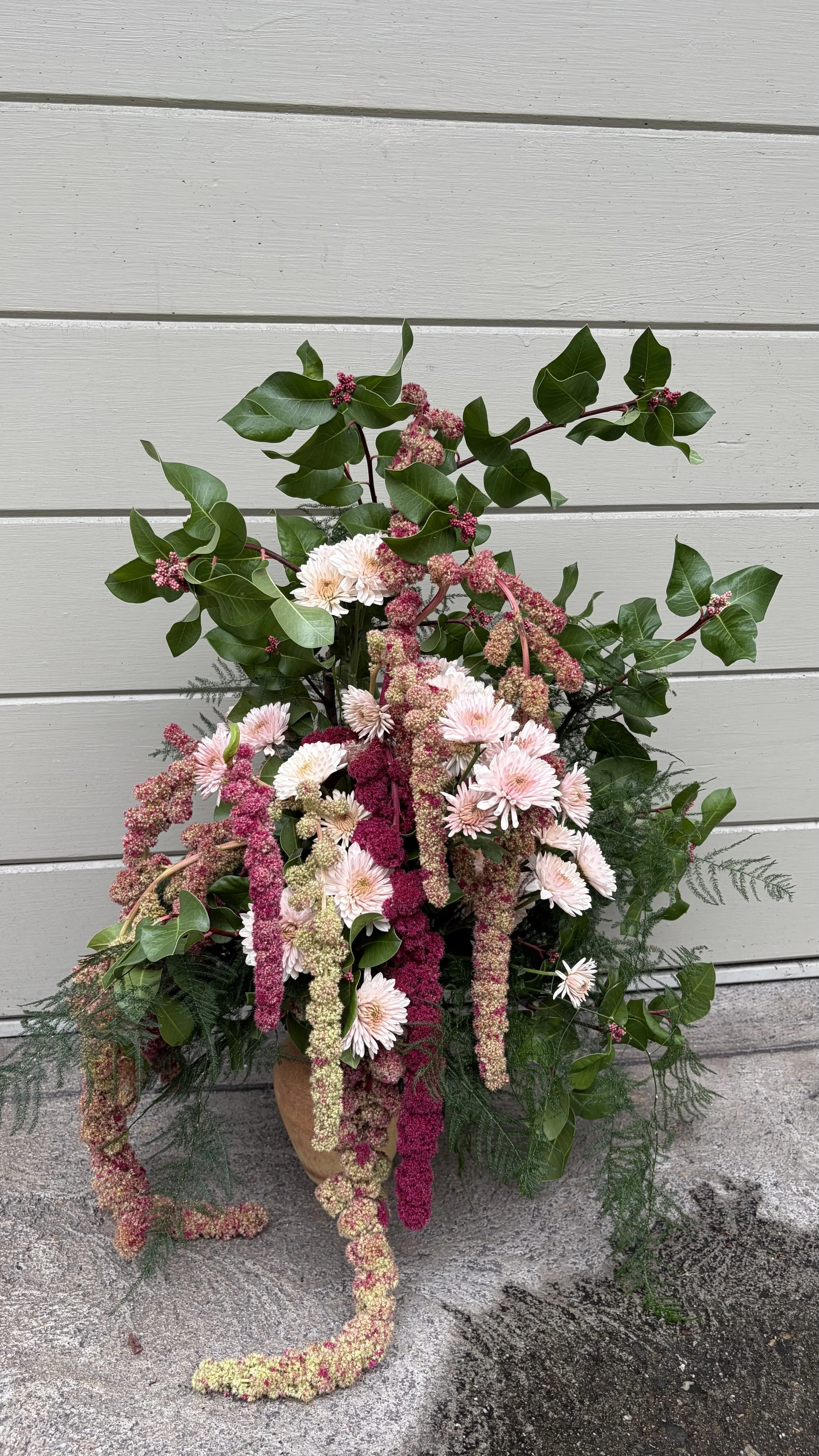 A flower arrangement in a ceramic pot featuring pink and white flowers with green foliage, placed on a concrete surface against a white horizontal wooden panel background.