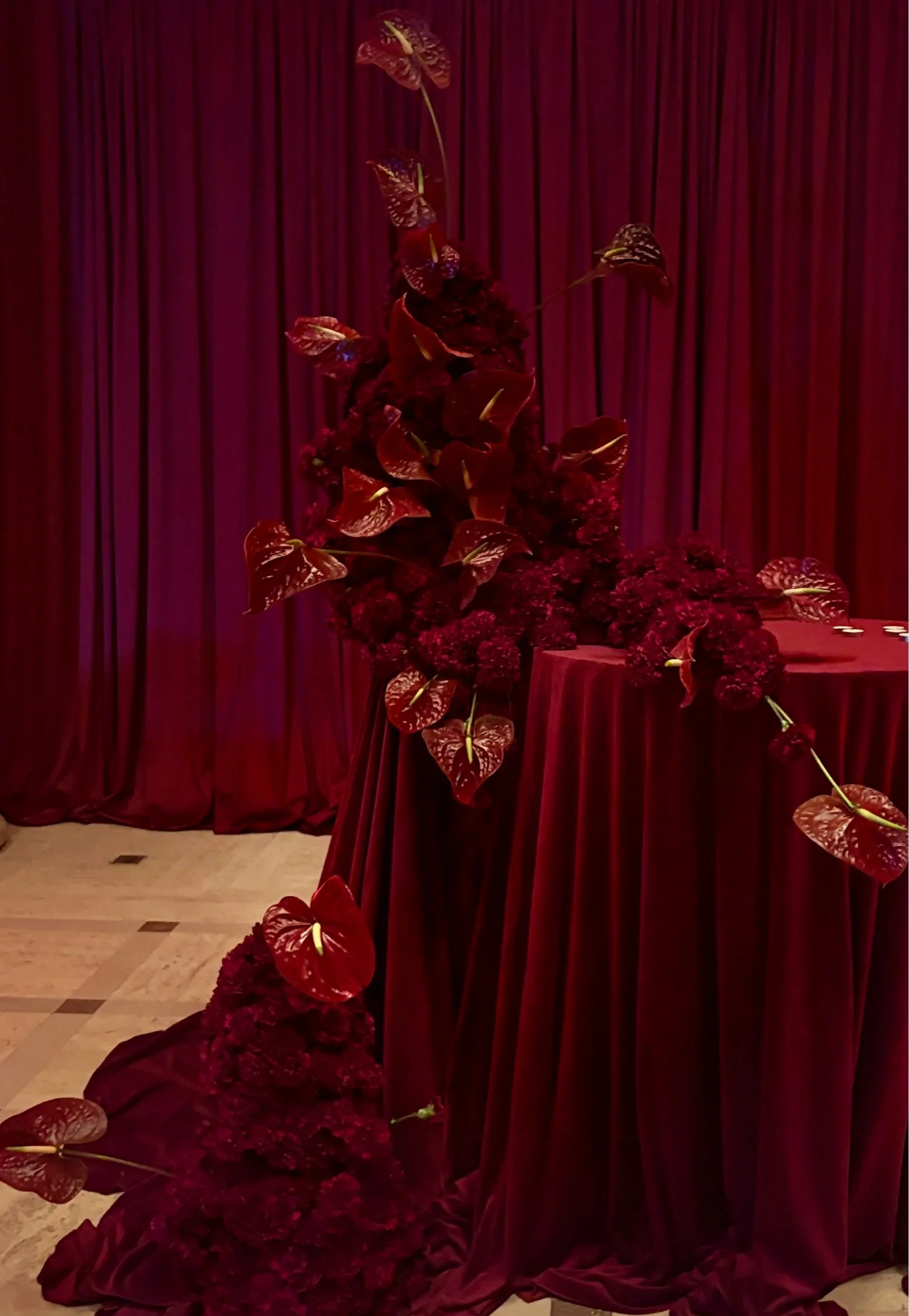 A floral arrangement of dark red flowers and red anthuriums on a table draped with red velvet fabric, with a matching backdrop of red velvet curtains.