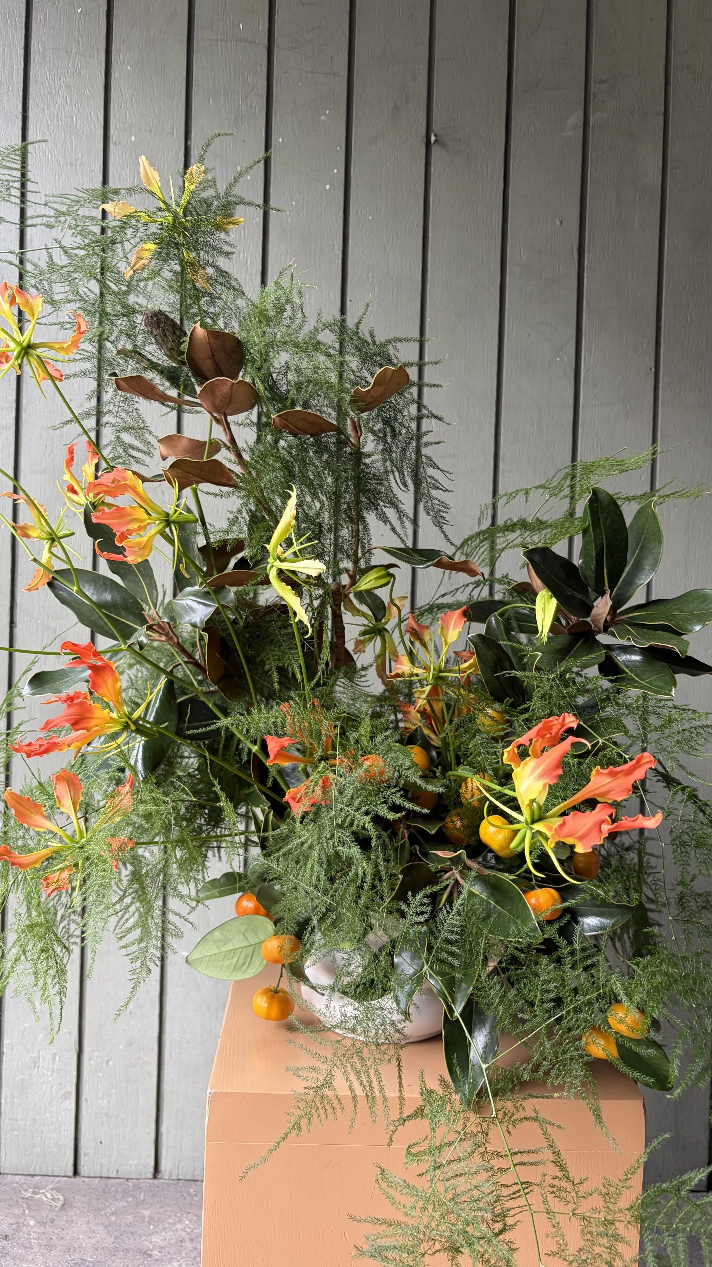 A floral arrangement with orange and yellow gloriosa lilies, green leaves, and orange berries, placed in a white pot on a peach-colored pedestal against a gray wooden wall.