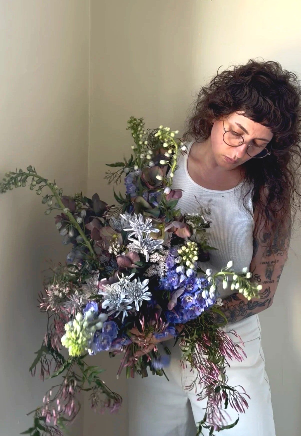 Woman with curly hair, glasses, and tattoos wearing a white sleeveless top holding a large bouquet of mixed flowers, standing against a beige wall.