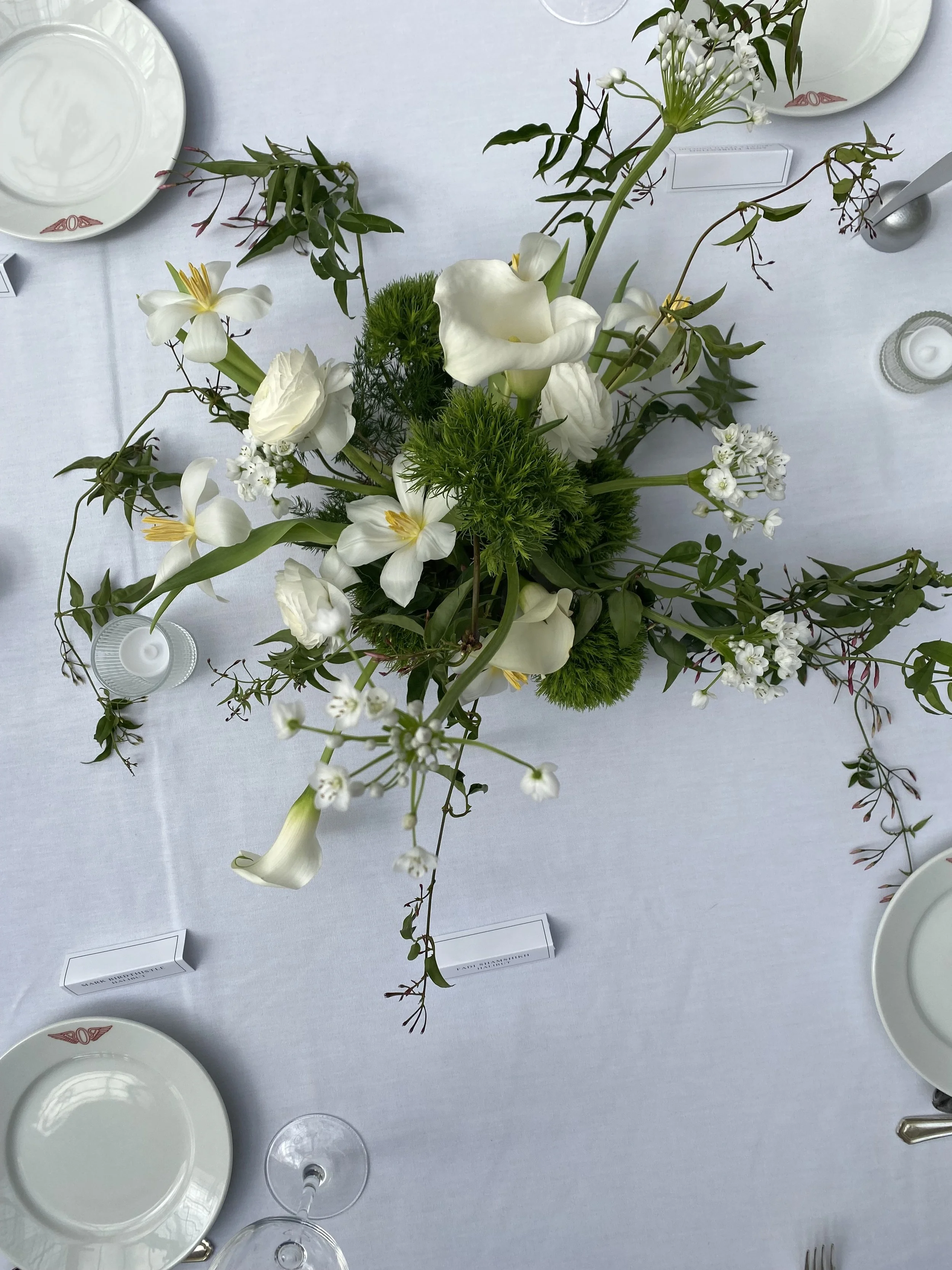 A top-down view of a floral centerpiece featuring white tulips and calla lilies and green foliage on a white table with plates, glasses, and candles at a wedding
