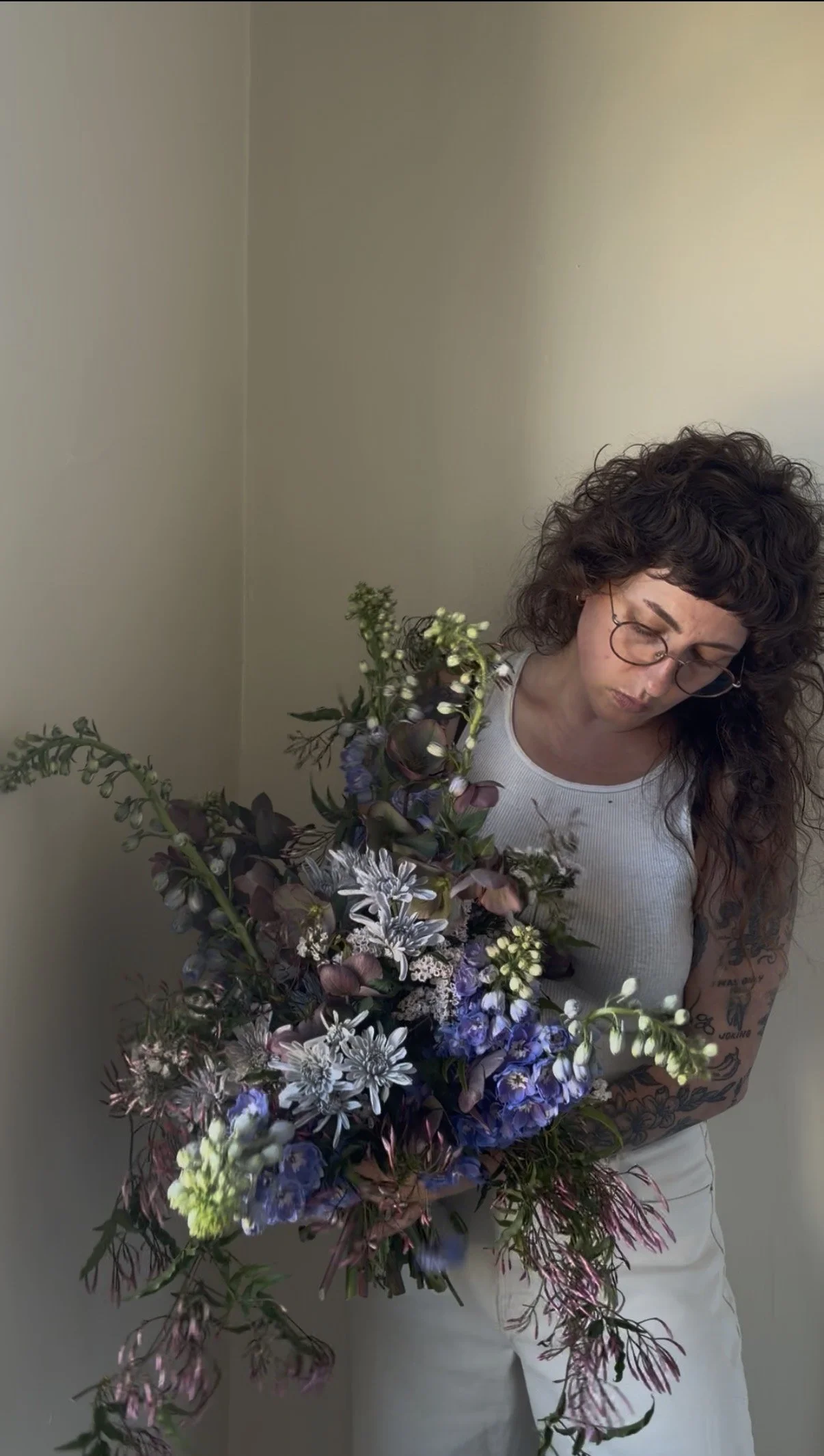 Woman with curly hair and glasses holding a large bouquet of purple, white, and green flowers.