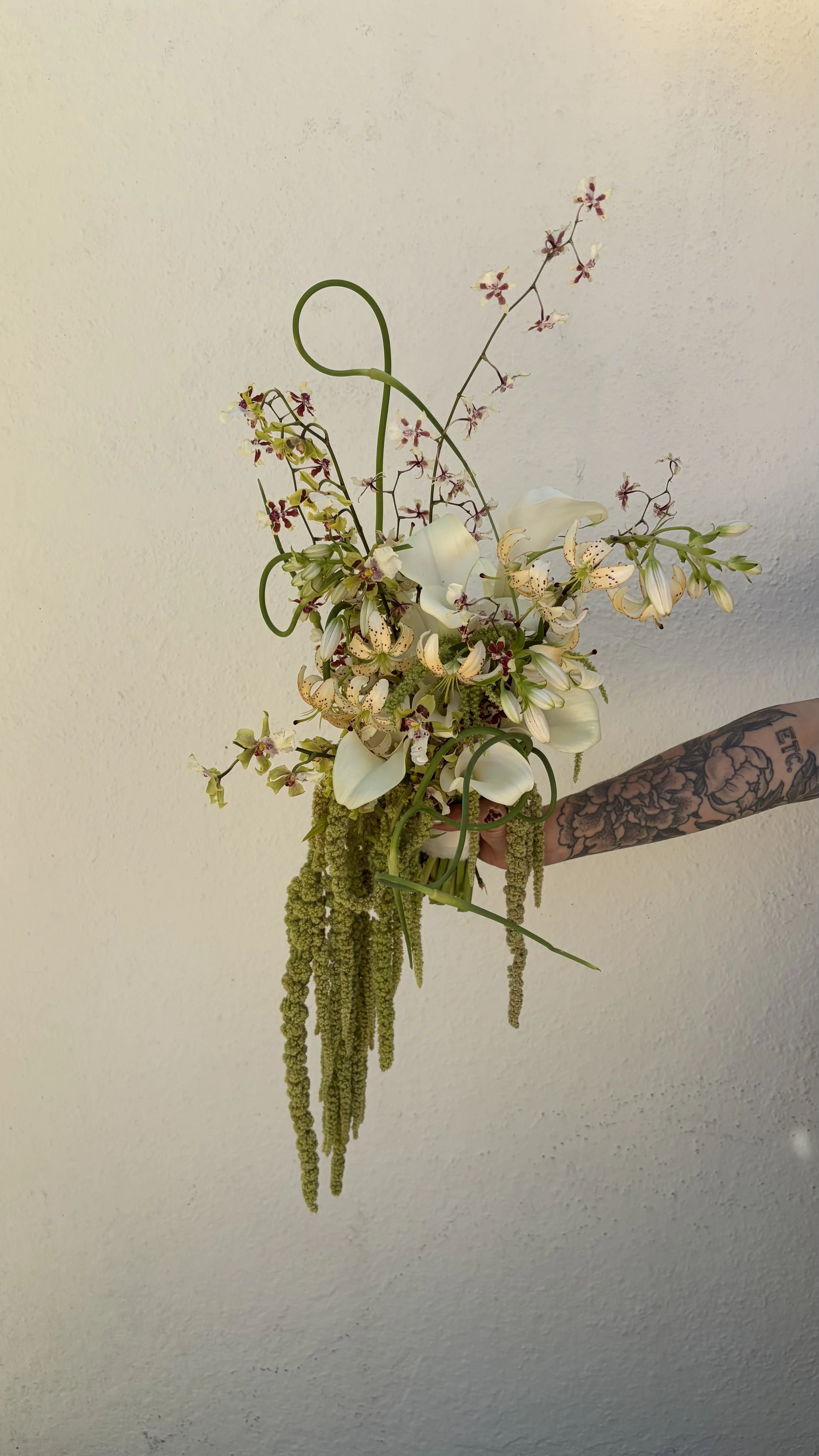 A person with a tattooed arm holding a bridal bouquet with calla lilies and amaranth against a plain wall background.