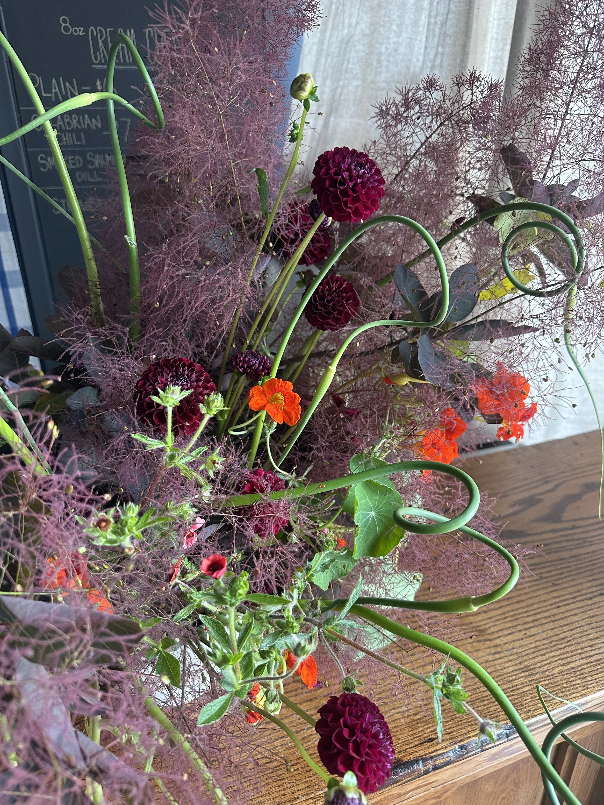 A vibrant floral arrangement featuring deep purple dahlias, bright orange flowers, and delicate pinkish-red feathery foliage on a wooden table.