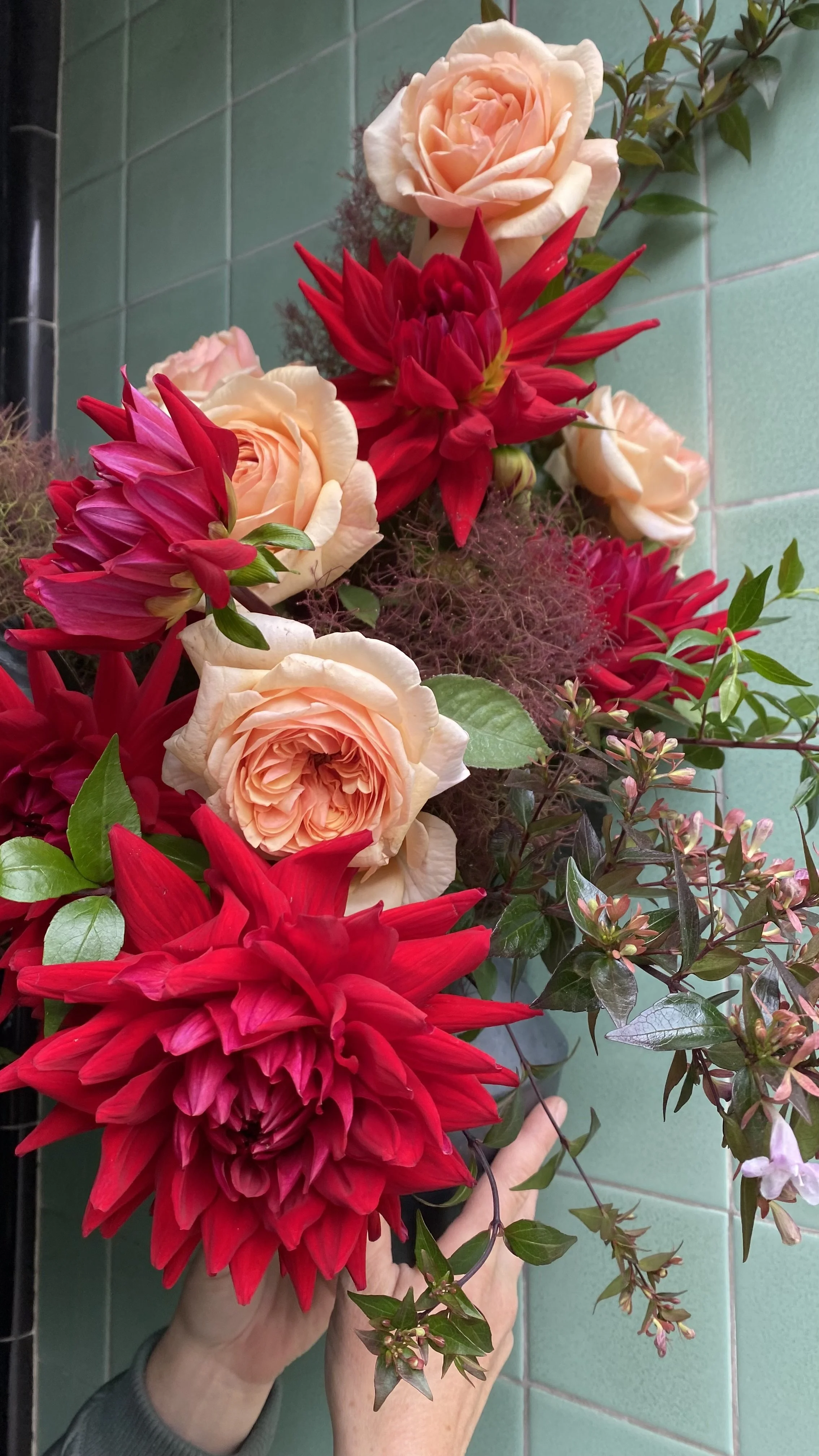 A person holding a colorful bouquet of large red dahlias, light pink roses, and green foliage against a tiled wall background.