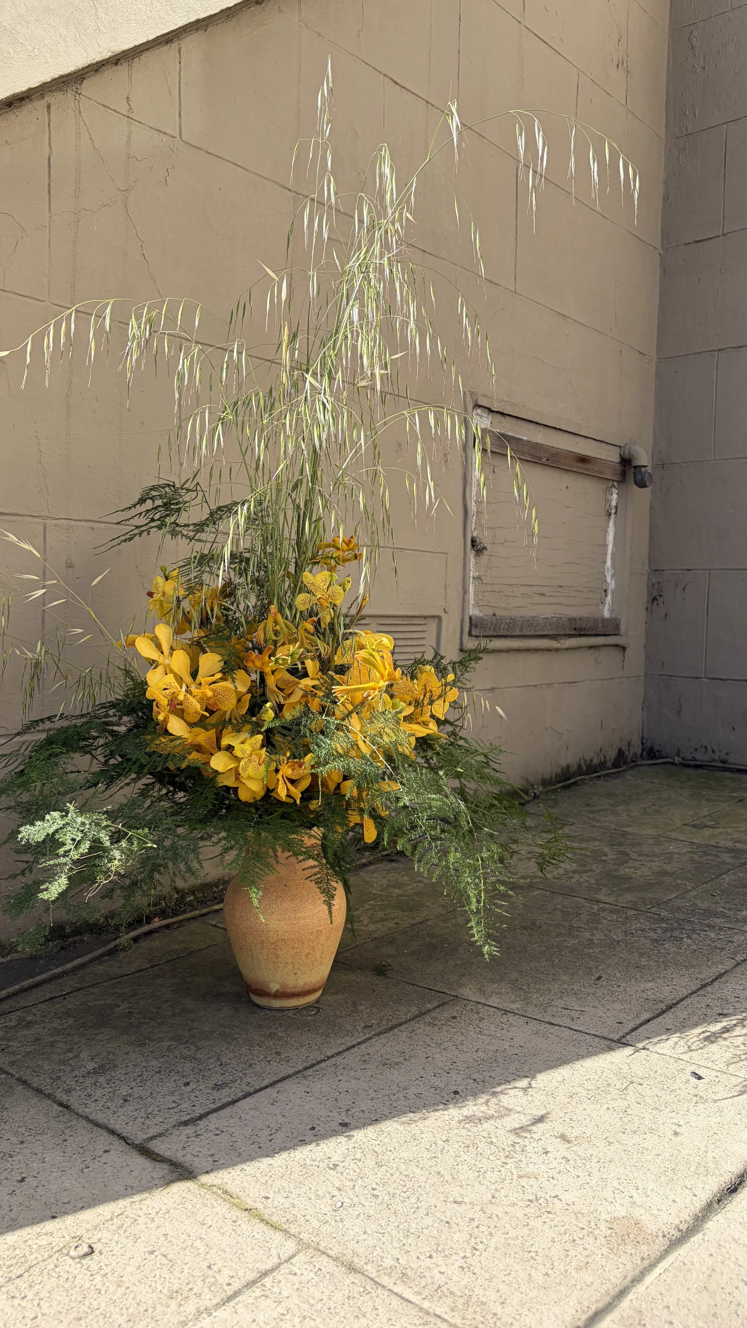 A flower arrangement in a brown vase placed on the sidewalk beside a beige wall with a small, boarded-up window.