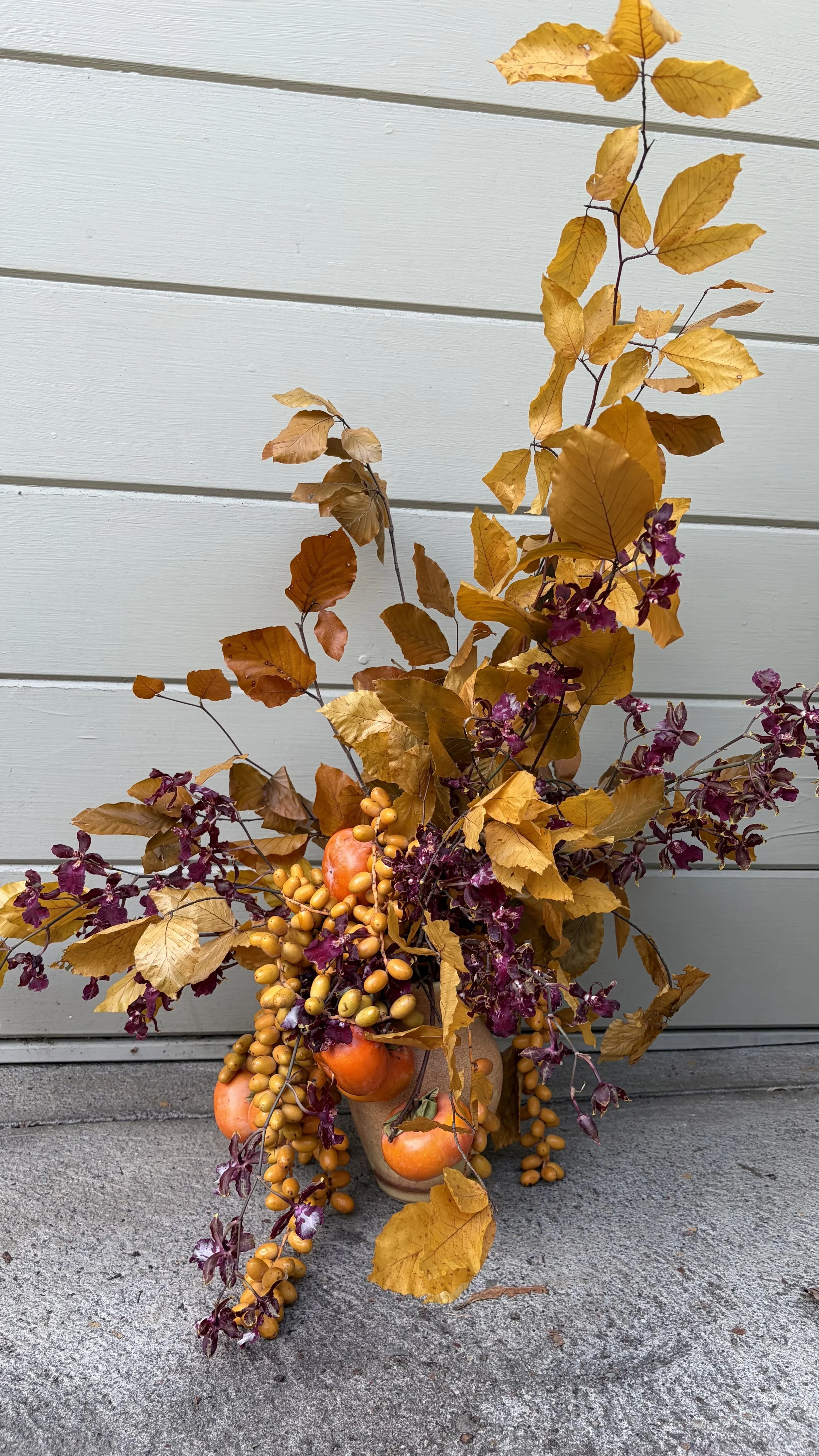 Autumnal floral arrangement with orange berries, persimmons, yellow and purple leaves in a vase, placed on concrete floor in front of a beige siding wall.