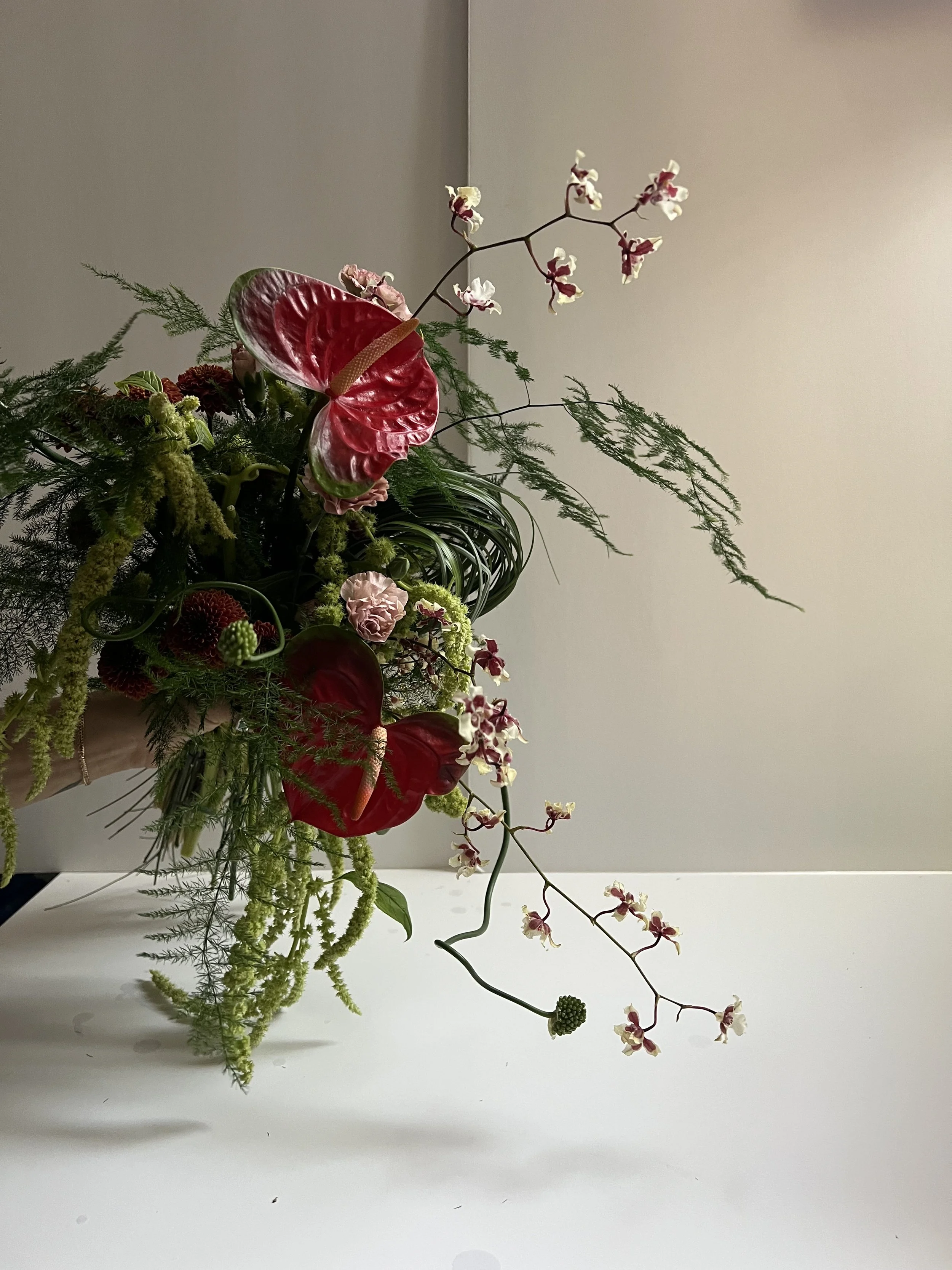 Flower arrangement including red anthuriums, orchids, green foliage, and hanging greenery in a vase.