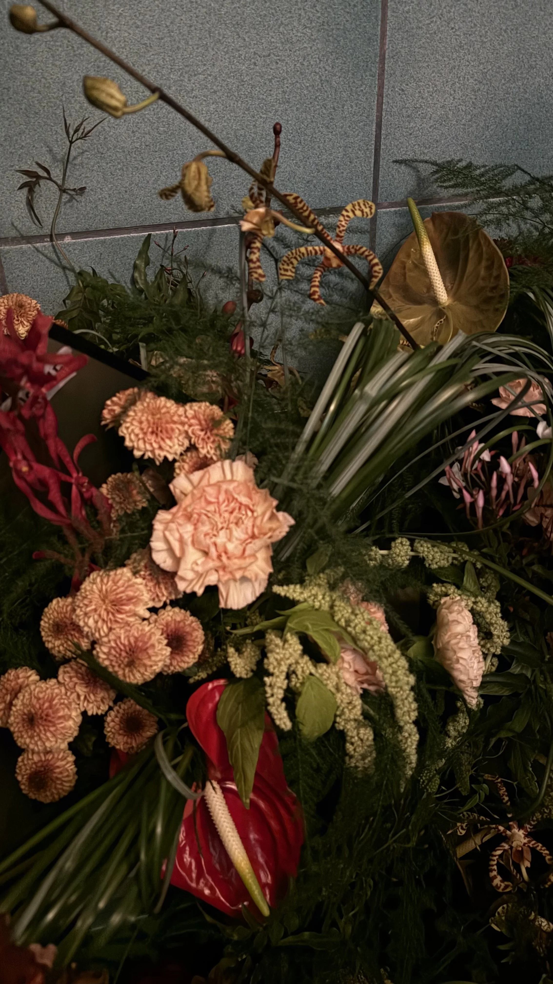 A floral arrangement with various flowers and greenery, including peach-colored carnations, white astilbe, and anthurium with a red spathe and yellow spadix, set against a grey tiled wall background.