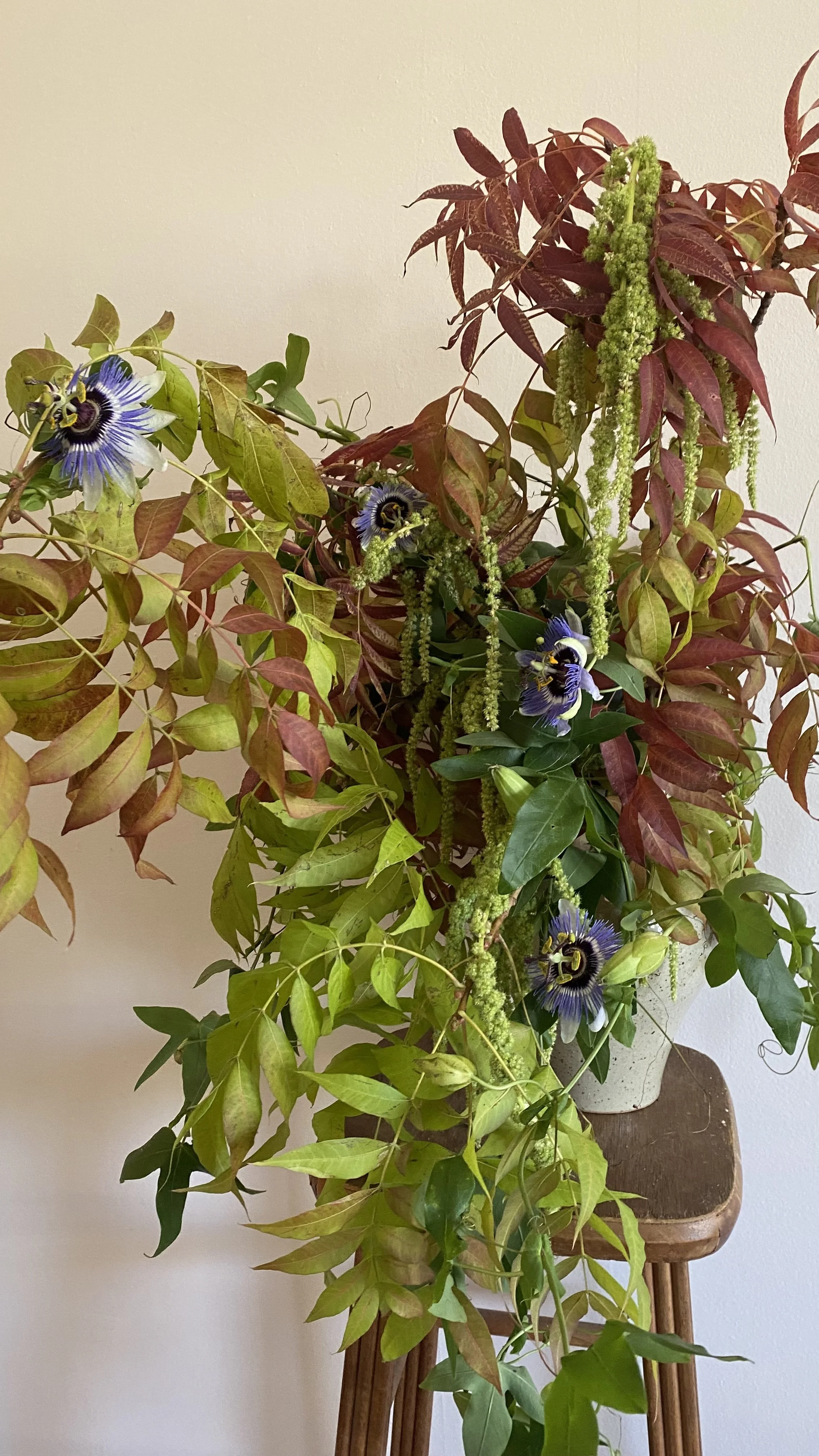 A lush floral arrangement in a white pot on a wooden stand, featuring green and red leaves along with purple and white passionflower flowers.