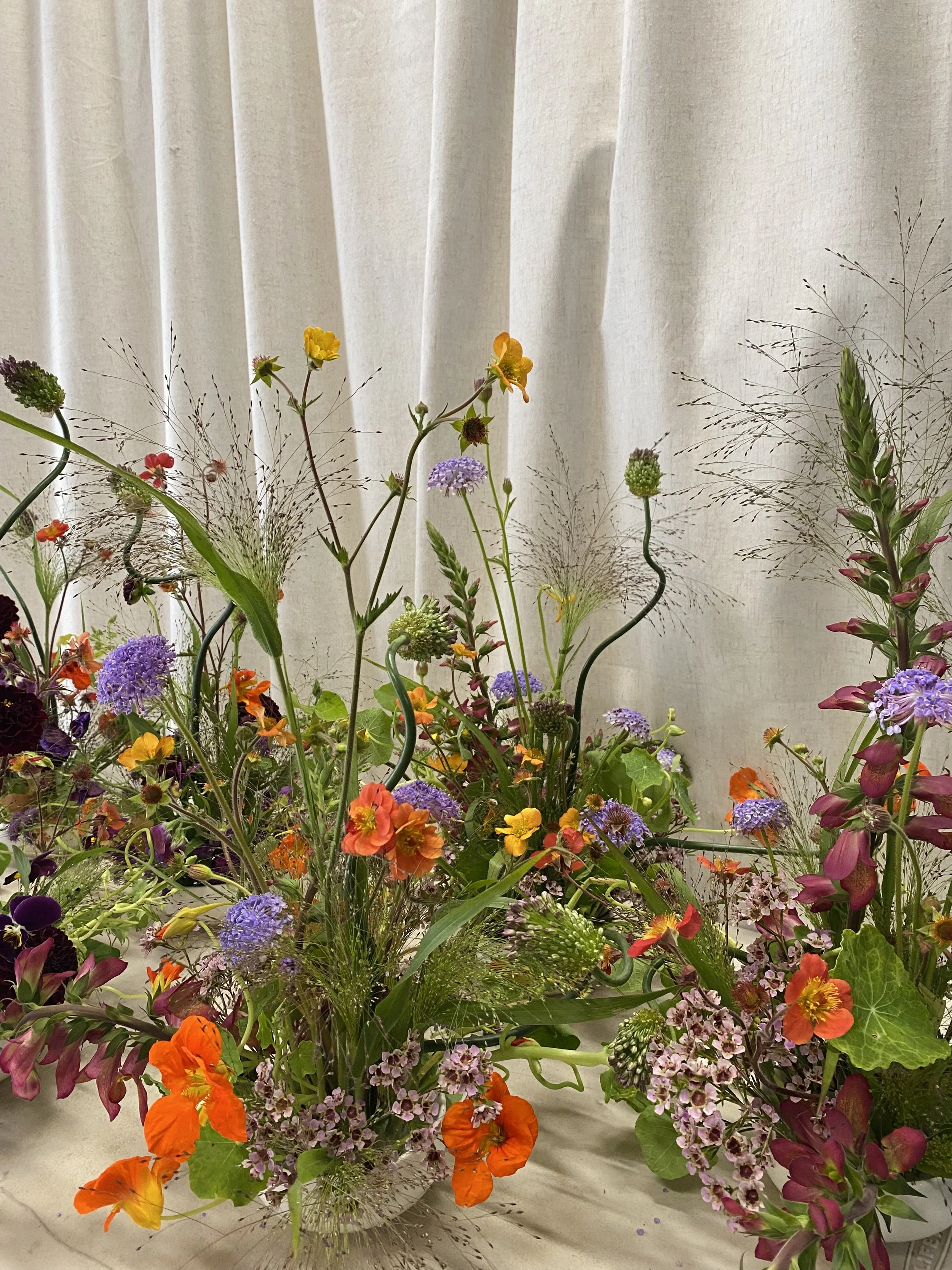 A colorful floral arrangement with various wildflowers in a glass vase, set against a white curtain background.