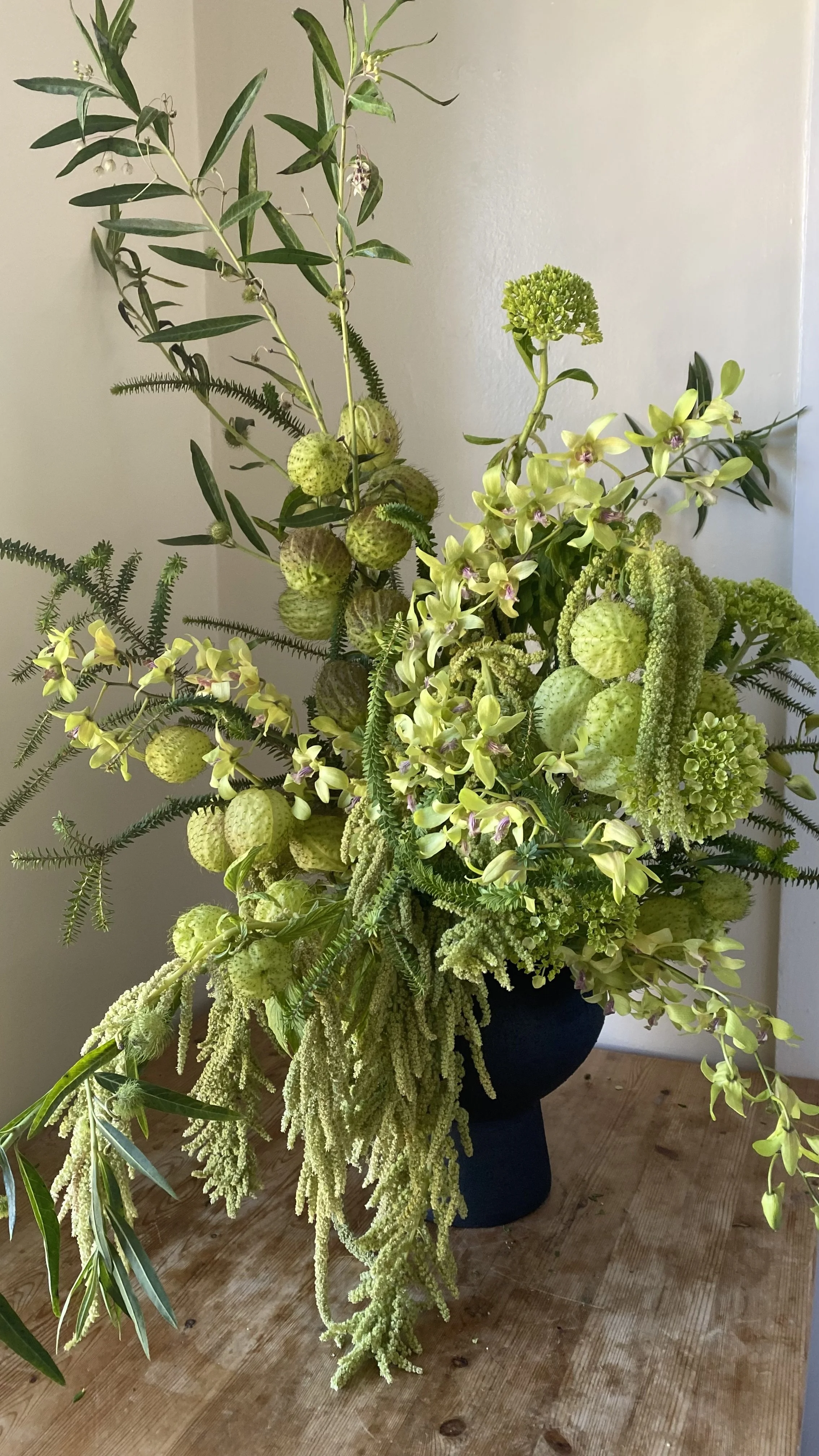 A floral arrangement with various green and yellow flowers and foliage in a black vase on a wooden surface.