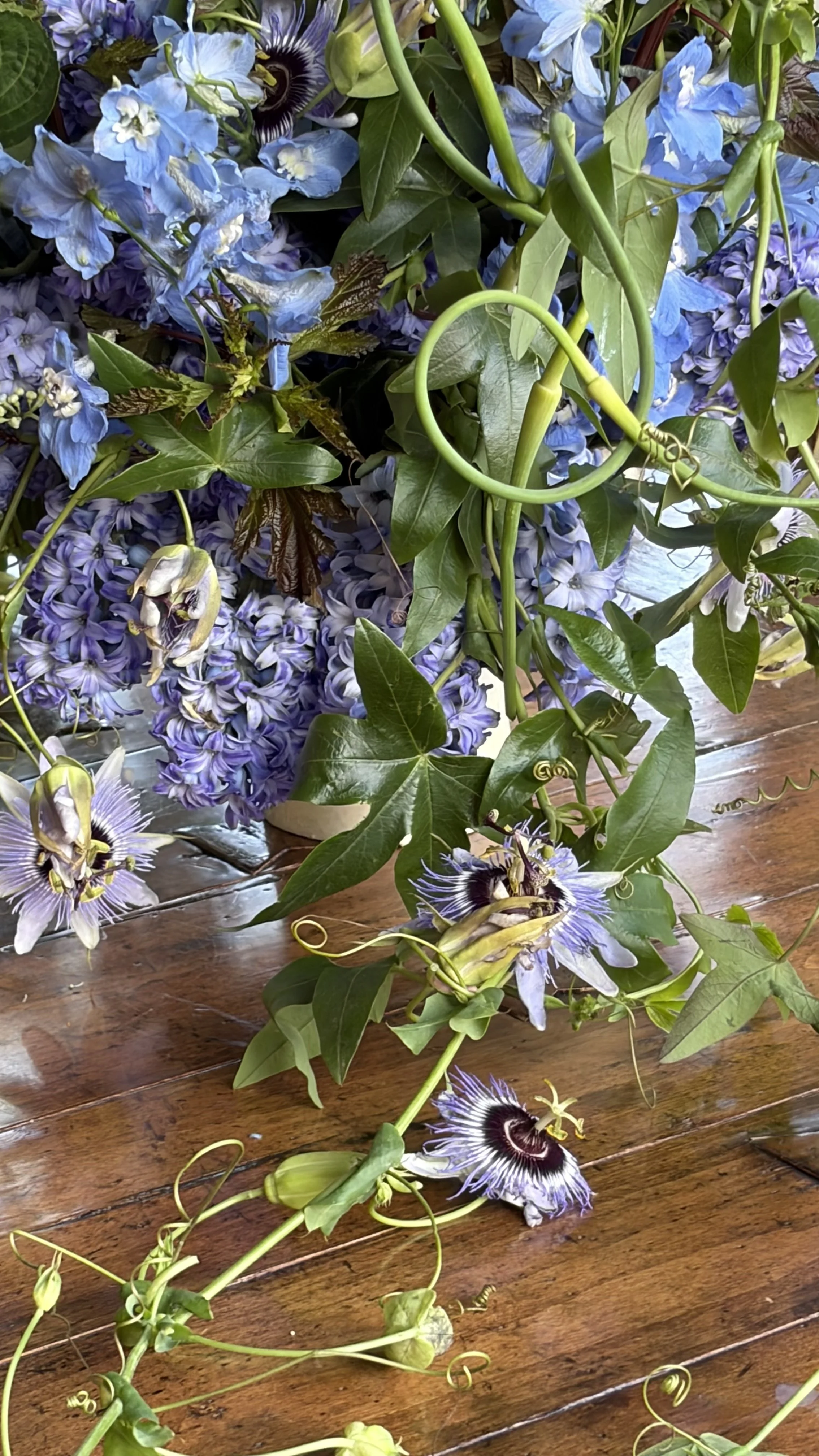 An arrangement of purple, blue, and white flowers with green leaves and vine tendrils on a wooden surface.