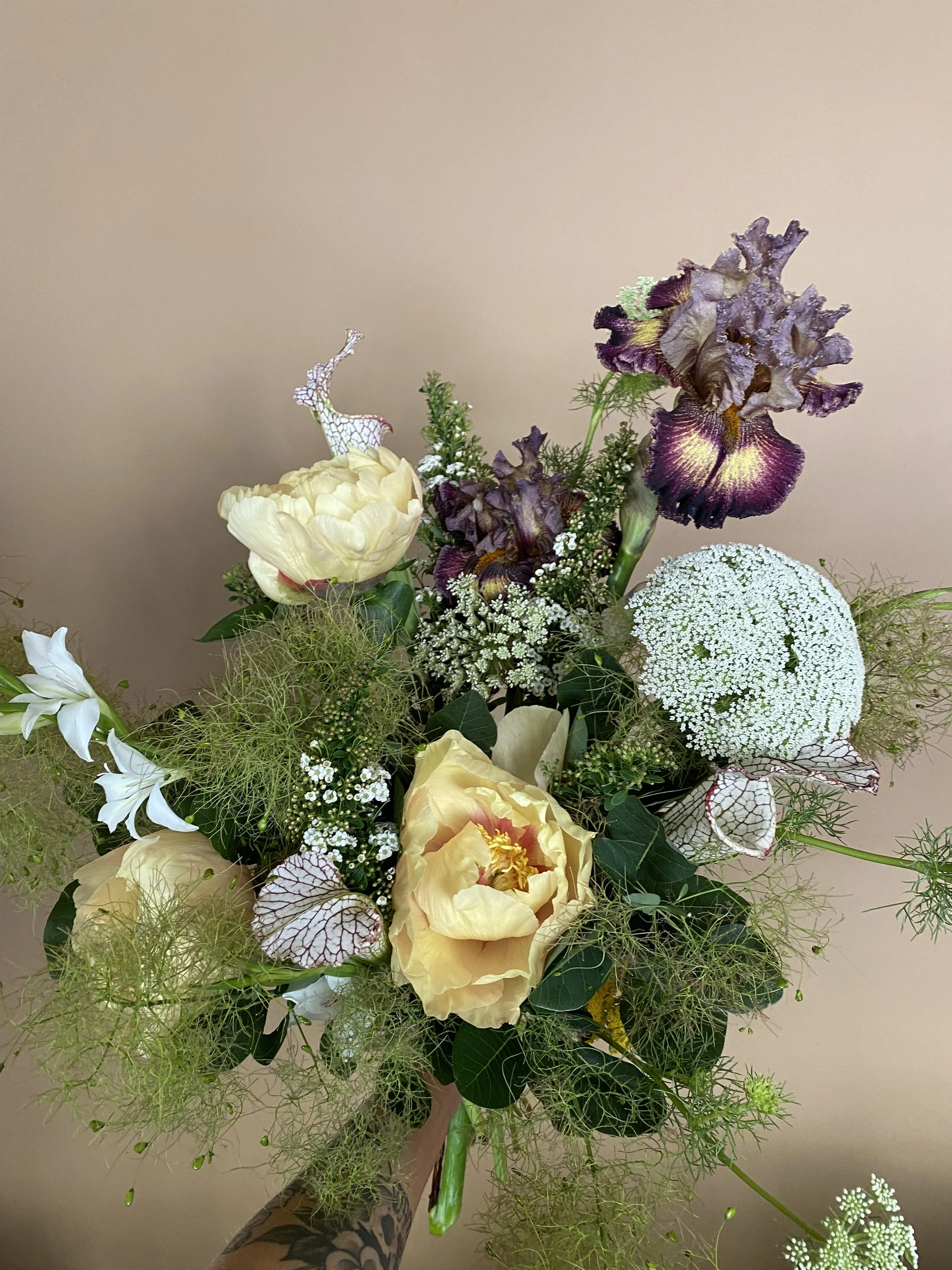 Arrangement of fresh flowers including white, yellow, and purple blooms with greenery and small white filler flowers, against a neutral background.