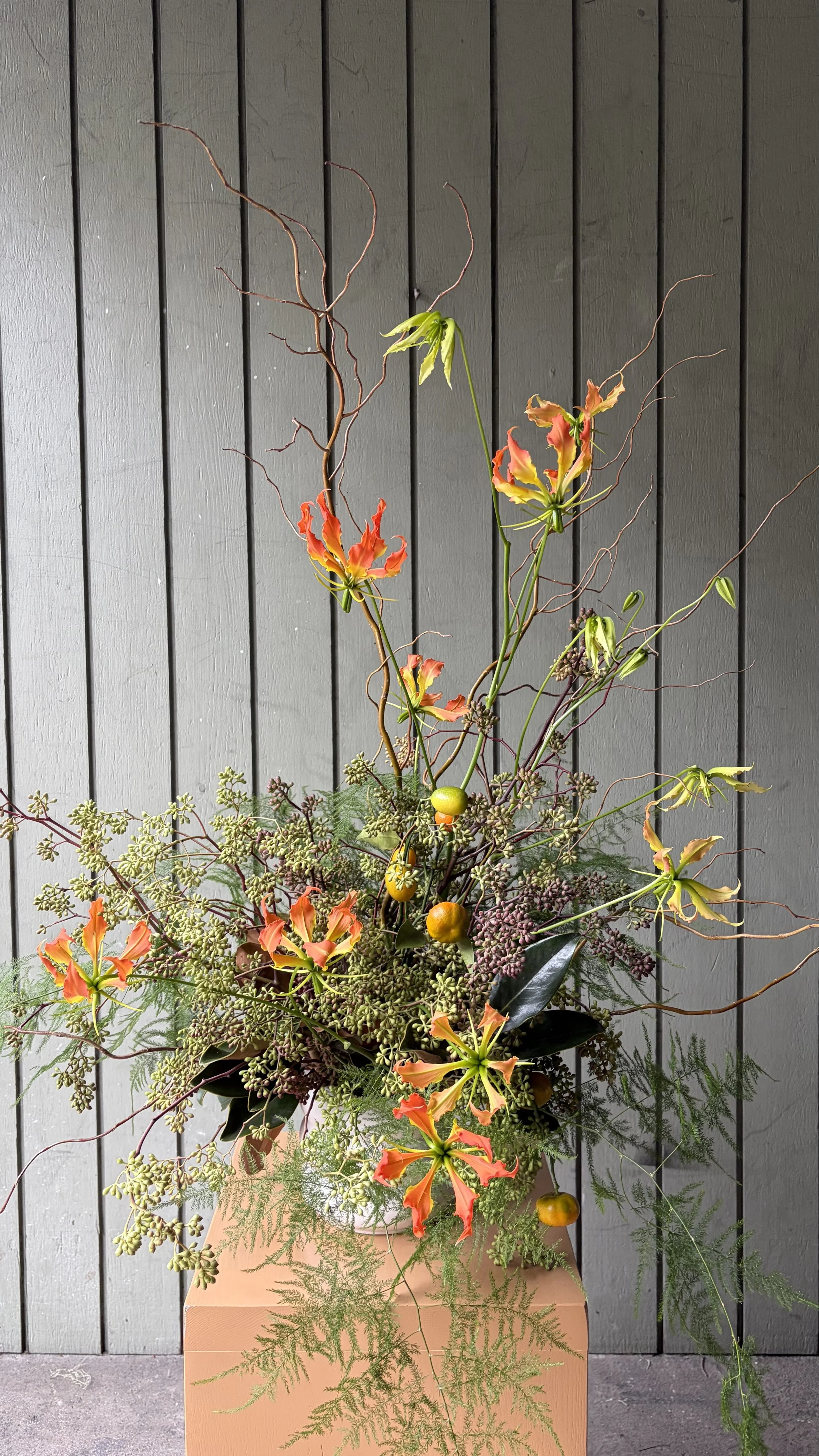 A tall floral arrangement featuring orange and yellow flowers, twisted branches, and green foliage in a light-colored vase placed on a peach-colored pedestal against a gray wood-paneled wall.